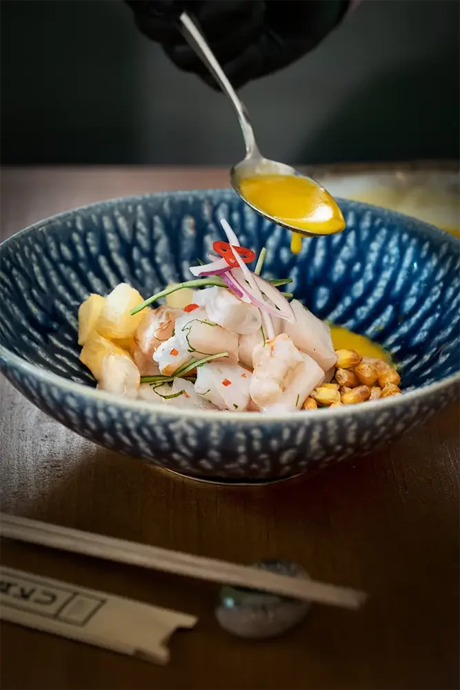En un tazón azul con textura, hay un platillo de ceviche con trozos de pescado, cebolla, limón y nueces, mientras que una mano vierte jugo de limón sobre el plato.
