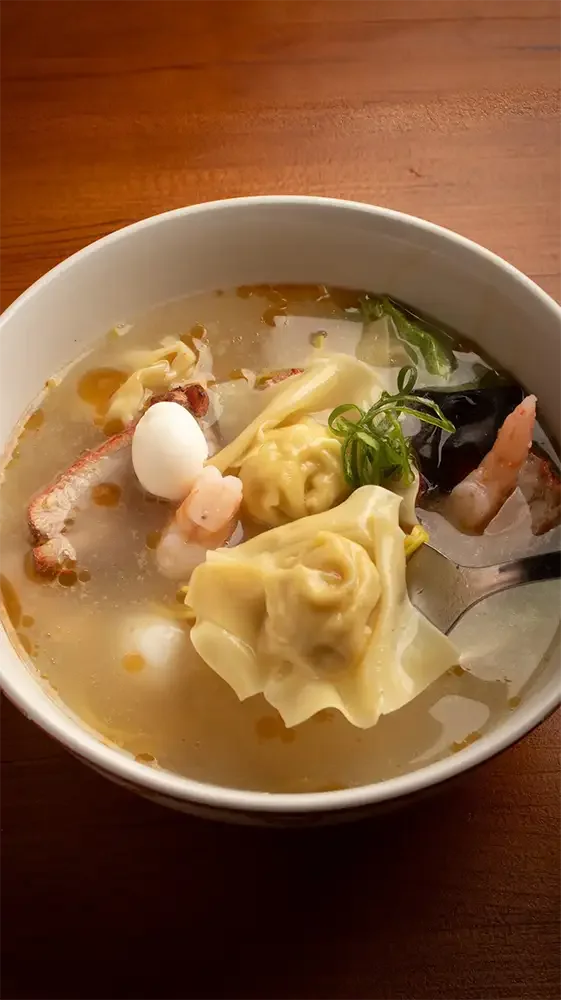 Sopa de wonton con ingredientes como wontons rellenos, carne de cerdo, vegetales y huevo en un tazón blanco.