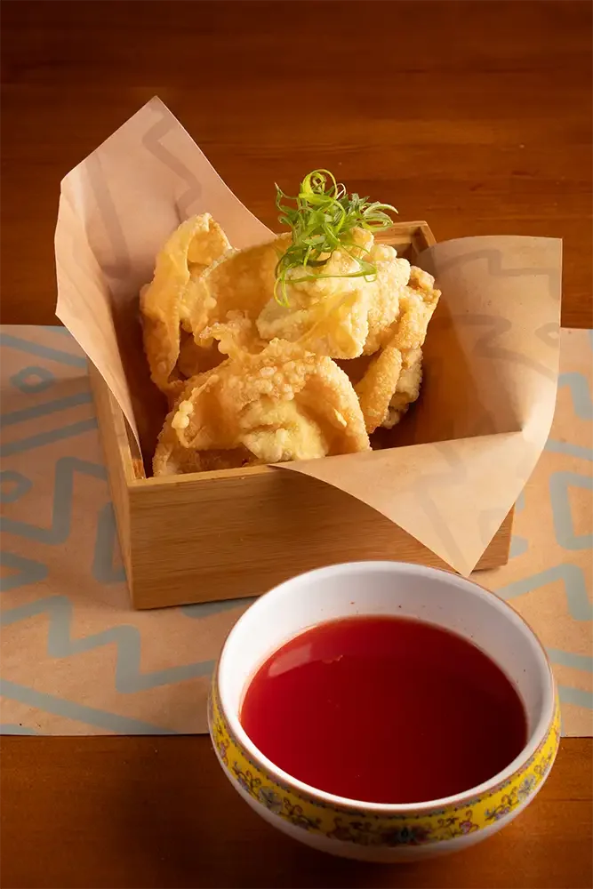 Piezas de calamar rebozadas y fritas en una bandeja de madera con adorno de hierba, acompañadas de una taza con salsa roja.