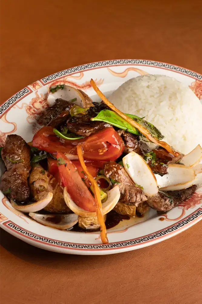 Plato de arroz, carne guisada con vegetales y salsa oscura, acompañado de tomates, cebollas y pimientos.