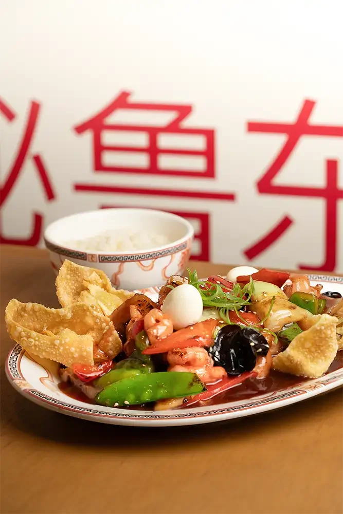 Plato de comida con verduras, mariscos, huevo y cortes de cerdo, acompañado de un recipiente con salsa, sobre una mesa de madera y fondo de pared con caracteres chinos en rojo.