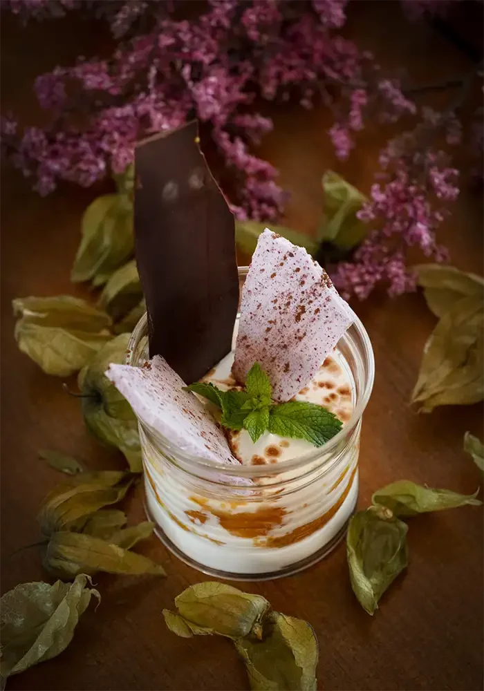 Postre en frasco con crema, galletas, hoja de menta y polvo de cacao, rodeado de flores secas y flores moradas en el fondo.