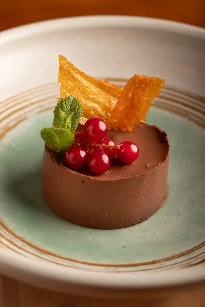 Postre de chocolate con hojas de menta y frutas rojas decorativas, acompañado de una galleta fina