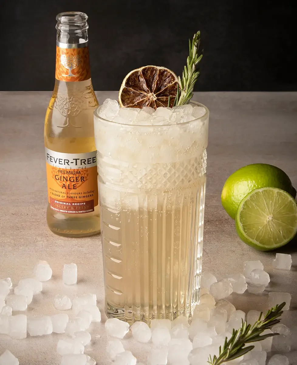 Vaso con bebida refrescante de color claro, lleno de hielo y decorado con una rodaja de limón seco y una ramita de romero, junto a un limón verde cortado y una botella de Ginger Ale, sobre una mesa con hielo y ramitas de romero.