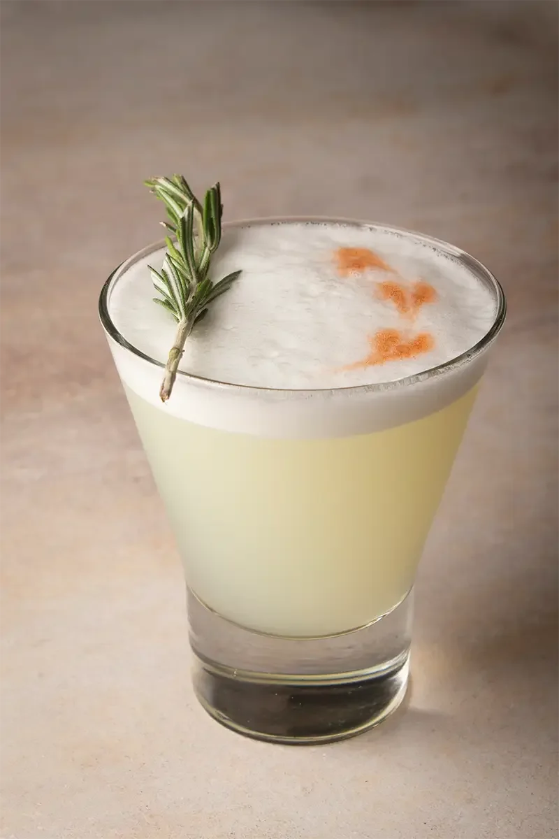 Cóctel con espuma blanca, decorado con una ramita de romero y un toque de polvo naranja en la parte superior, en un vaso transparente