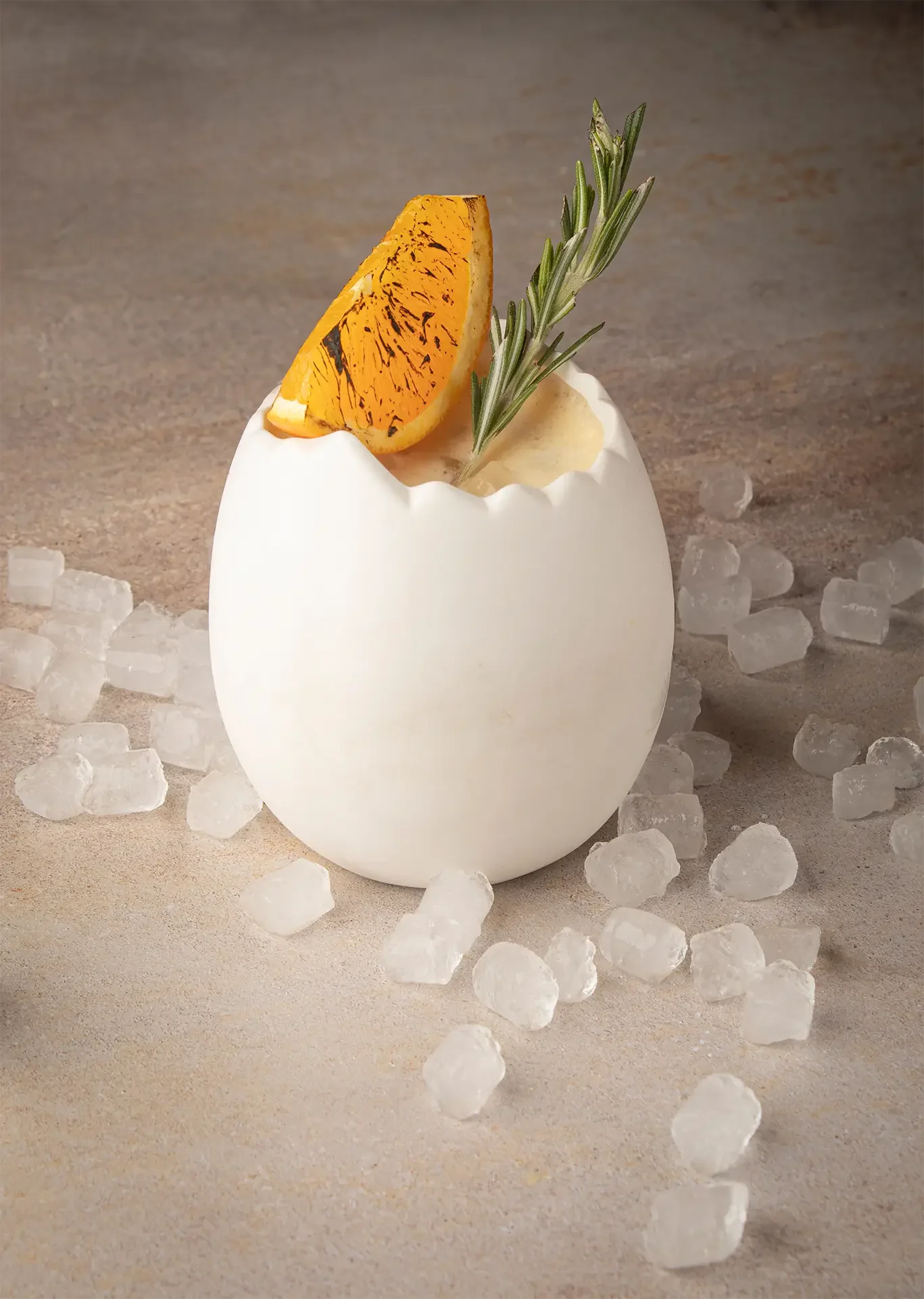 Huevo blanco roto con una rebanada de naranja y una ramita de romero en su interior, rodeado de hielo en una superficie de arena fina.