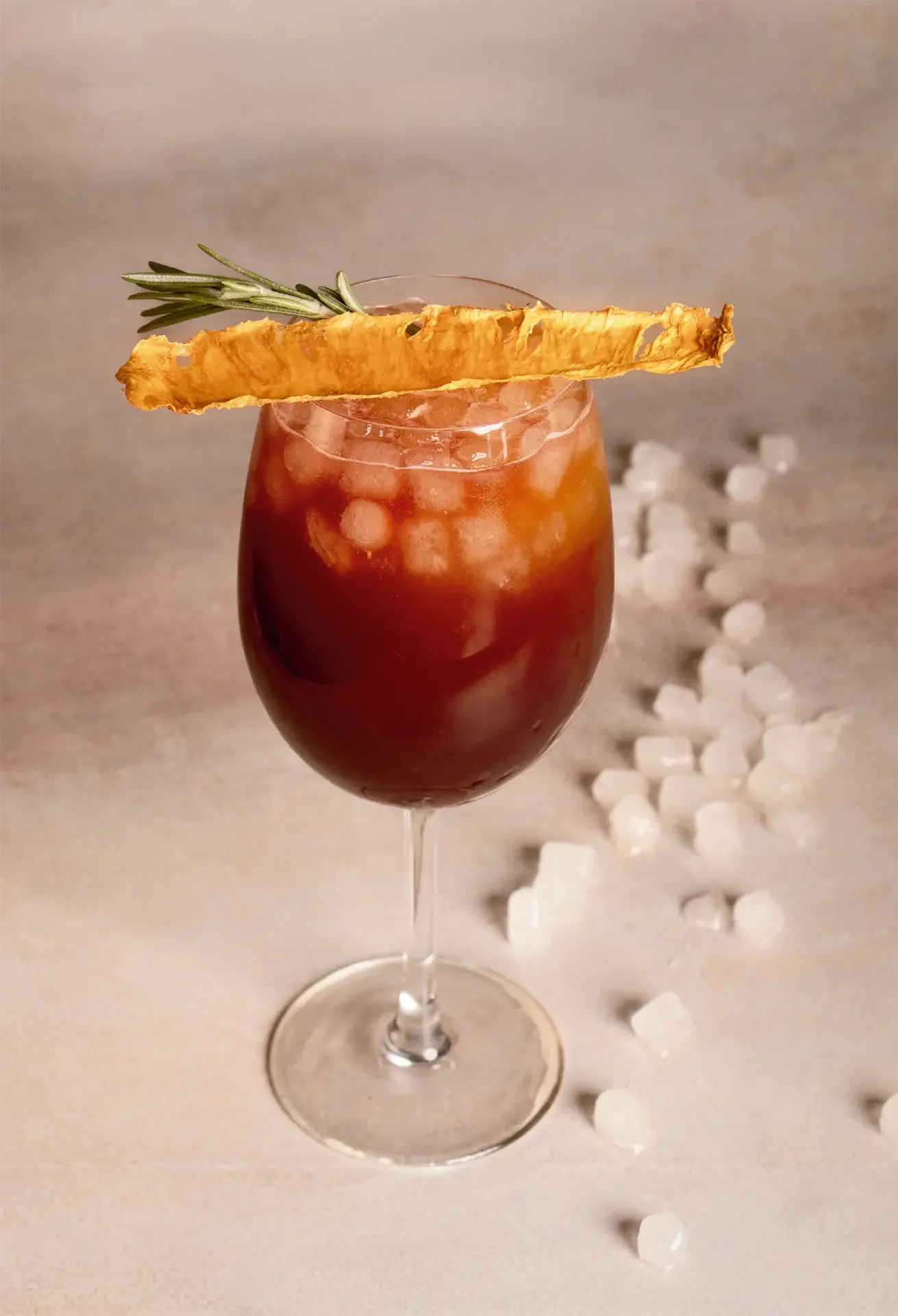 Cóctel rojo en copa de vino con hielo y una ramita de romero y cáscara de piña en el borde, rodeado de cubos de hielo en la mesa.
