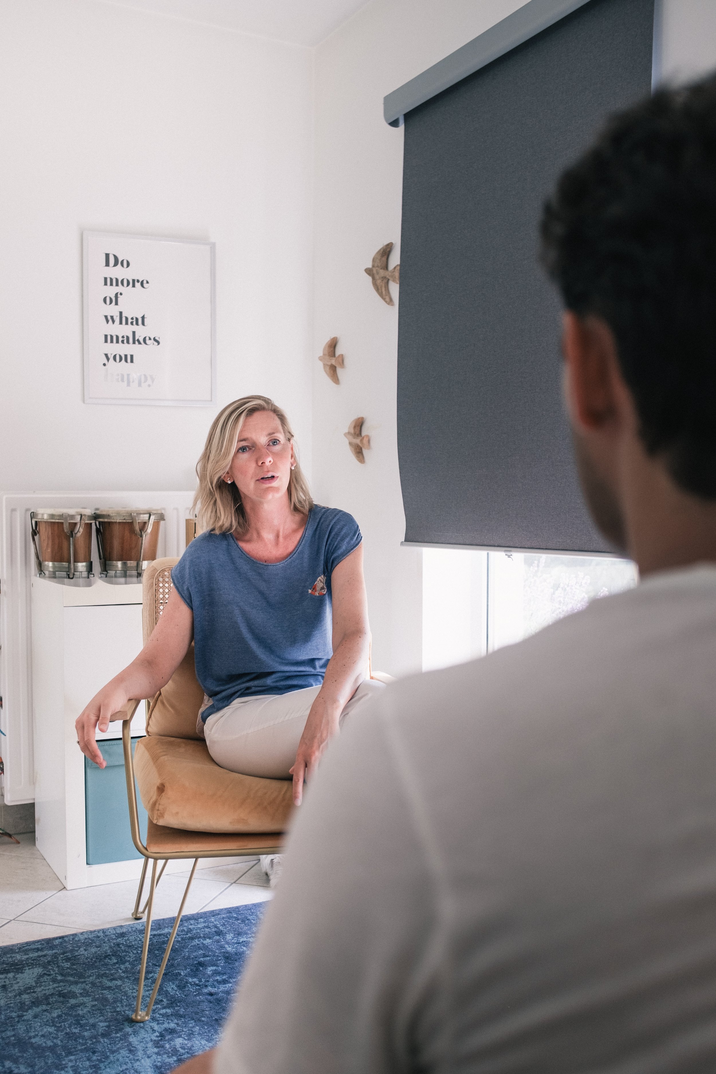 Une femme assise sur un fauteuil en cuir beige, en conversation avec un homme dont on ne voit pas le visage, dans une pièce lumineuse avec décoration murale de oiseaux en bois et un panneau avec une citation.