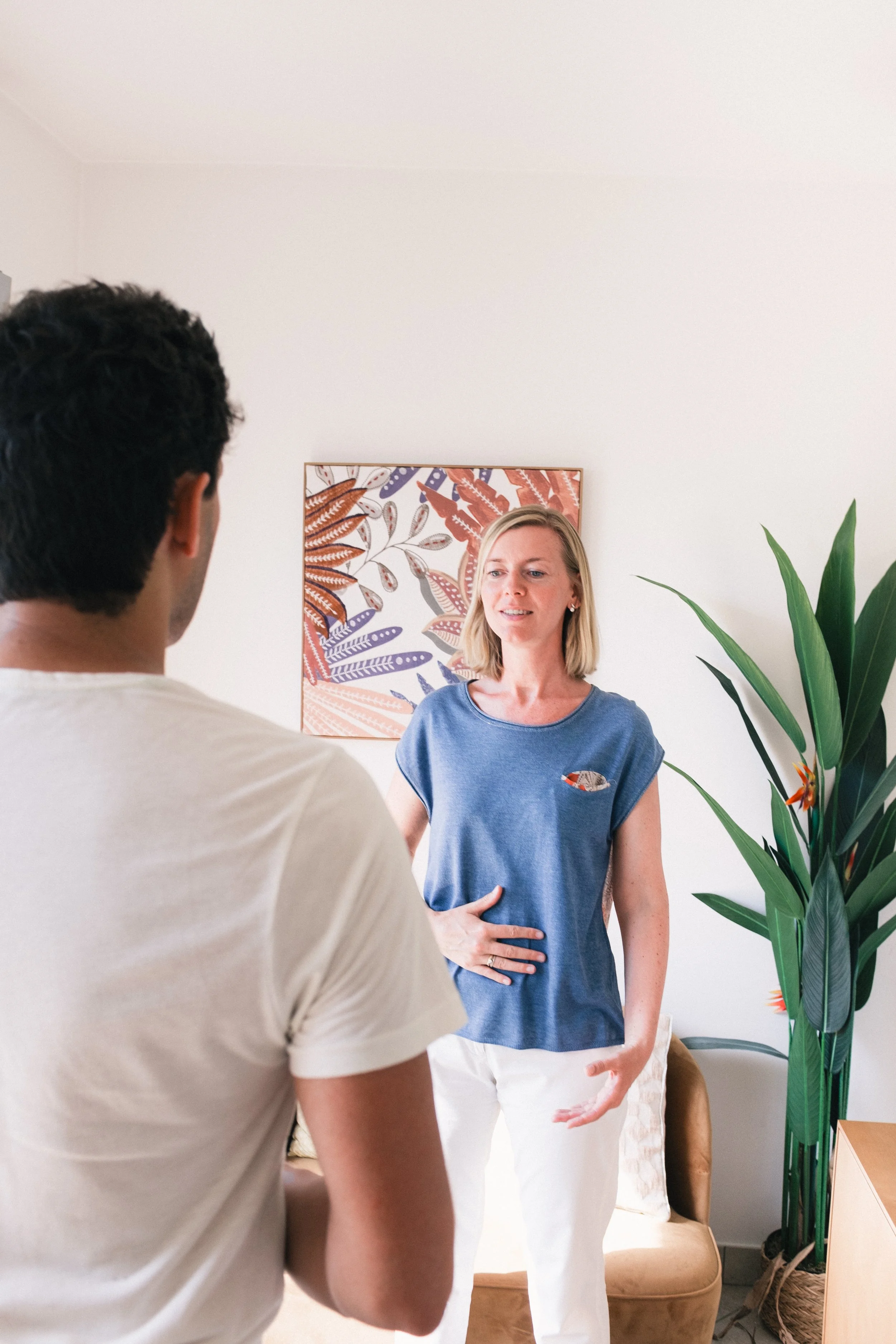 Une femme avec les cheveux blonds, portant un t-shirt bleu, semble expliquer quelque chose à un homme, vu de dos, qui porte un t-shirt blanc. La scène se déroule dans un intérieur lumineux avec un tableau coloré derrière la femme, une grande plante verte à droite et un fauteuil beige au fond.