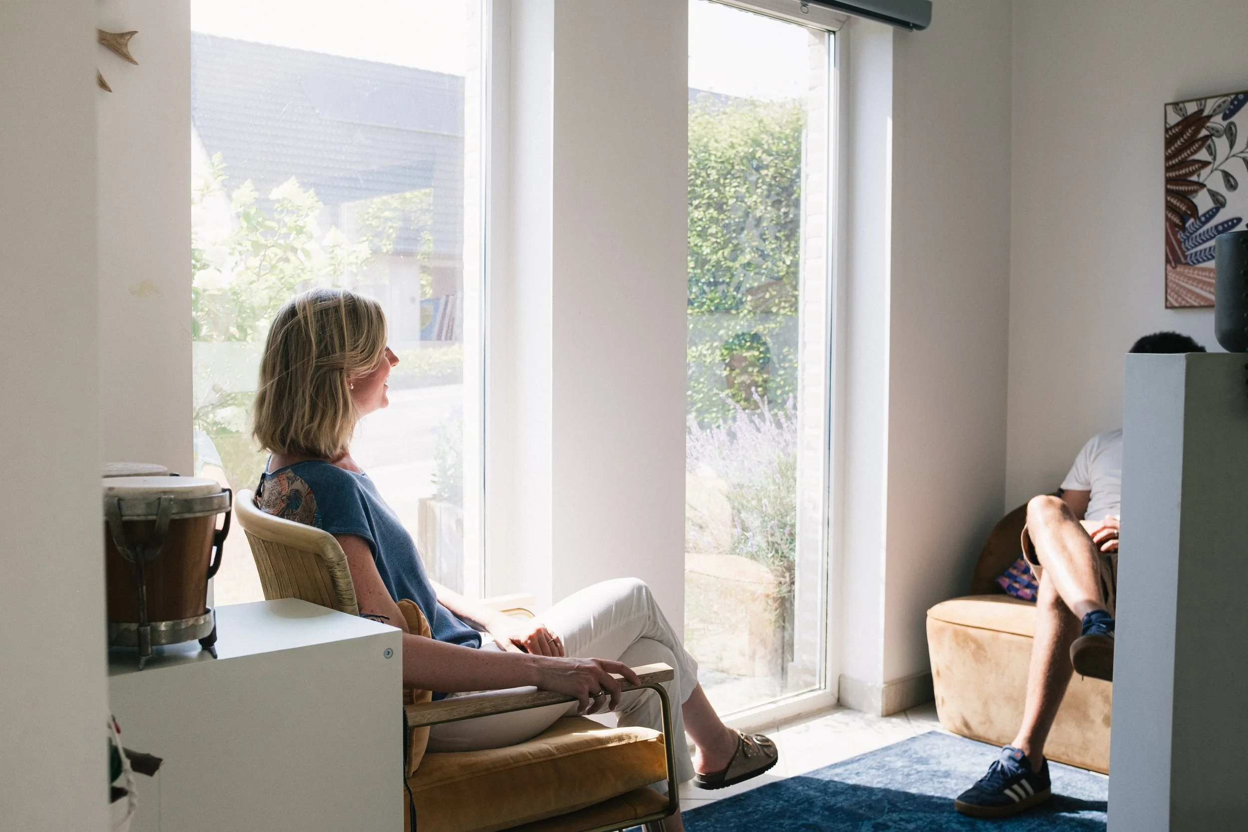 Une femme assise sur une chaise à l'intérieur d'une maison, regardant par une grande fenêtre avec vue sur un jardin. Deux autres personnes sont partiellement visibles dans la pièce.
