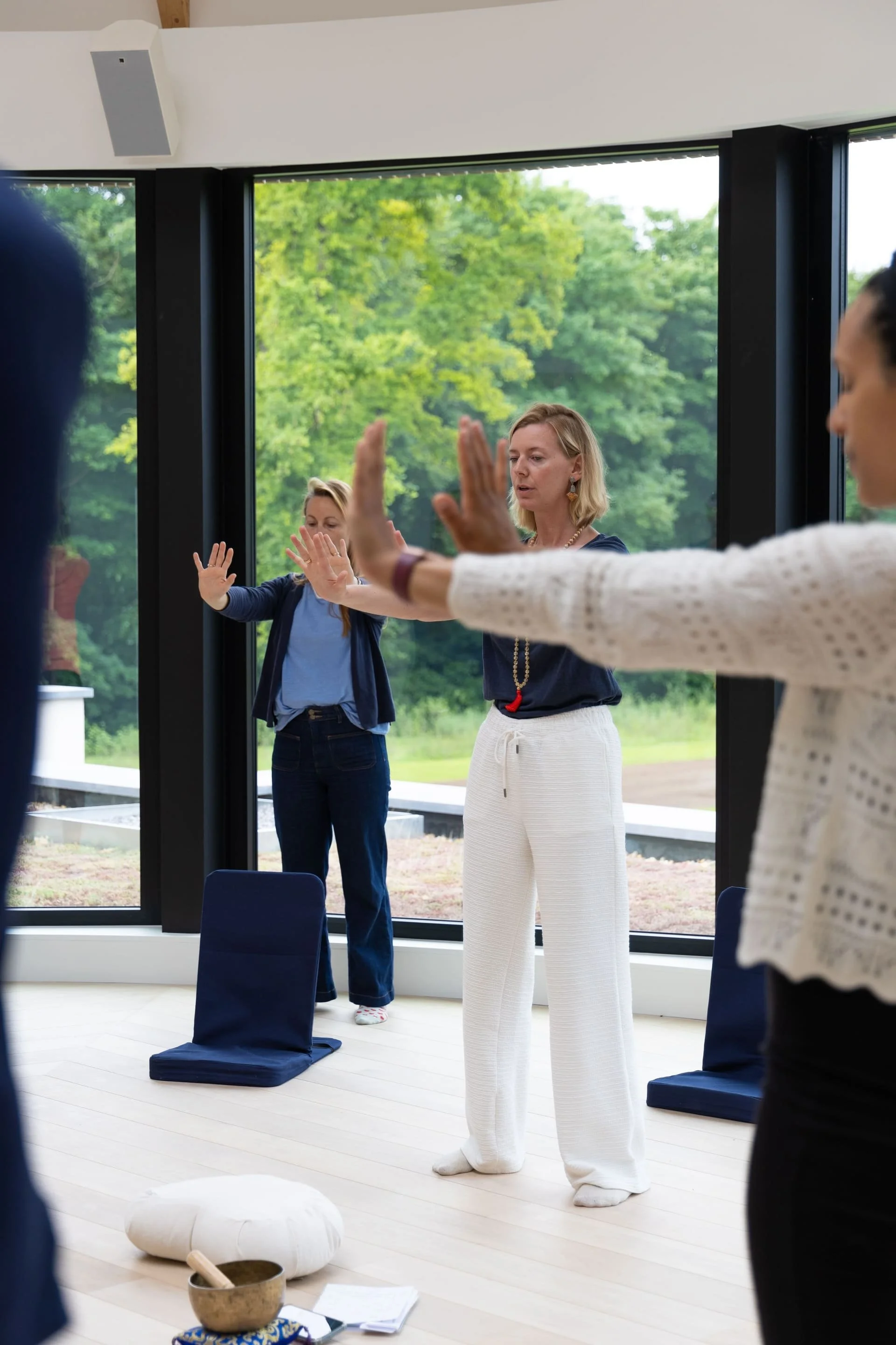 Groupe de personnes pratiquant une séance de méditation ou de relaxation dans une salle lumineuse avec de grandes fenêtres. Certaines personnes ont les mains levées en position de méditation. Il y a un tapis de méditation, un bol en métal, une pierre et des papiers sur le sol.