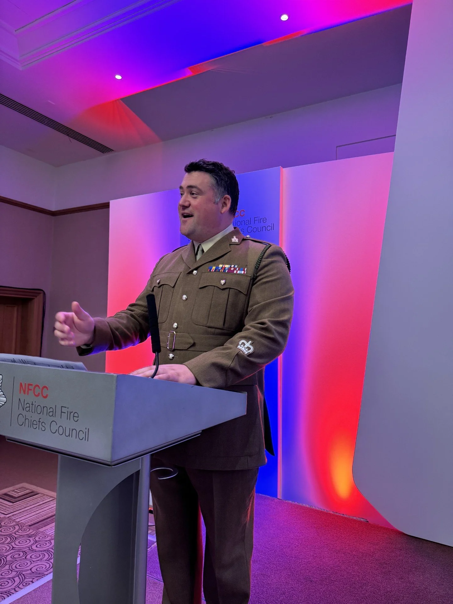 A man in a military uniform standing at a podium, speaking at the NFCC National Fire Chiefs Council event, with multicolored pink, purple, and red lighting in the background.