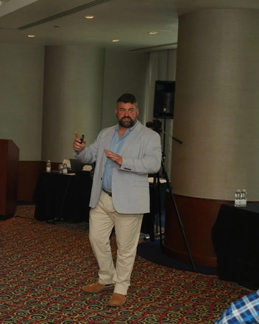 Man giving a presentation or speech in a conference room, holding a remote control, dressed in a light blazer, blue shirt, beige pants, and brown shoes, with audio-visual equipment and water bottles in the background.