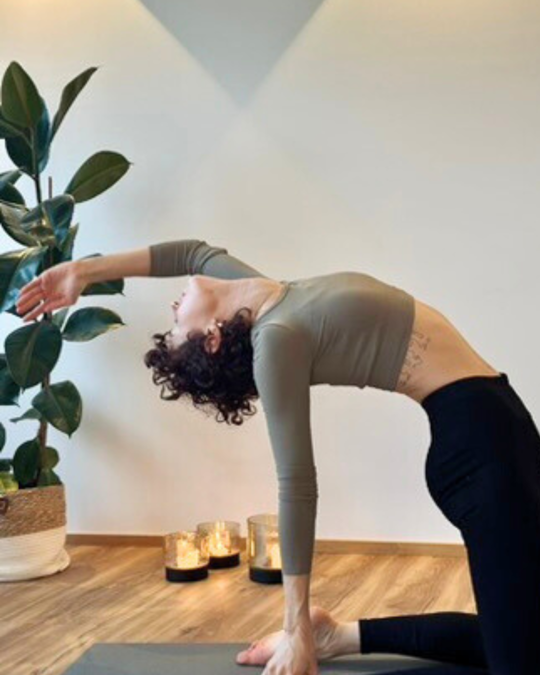 Frau macht Yoga in einer Wohnung, neben einer großen Pflanze und Kerzen auf dem Boden.
