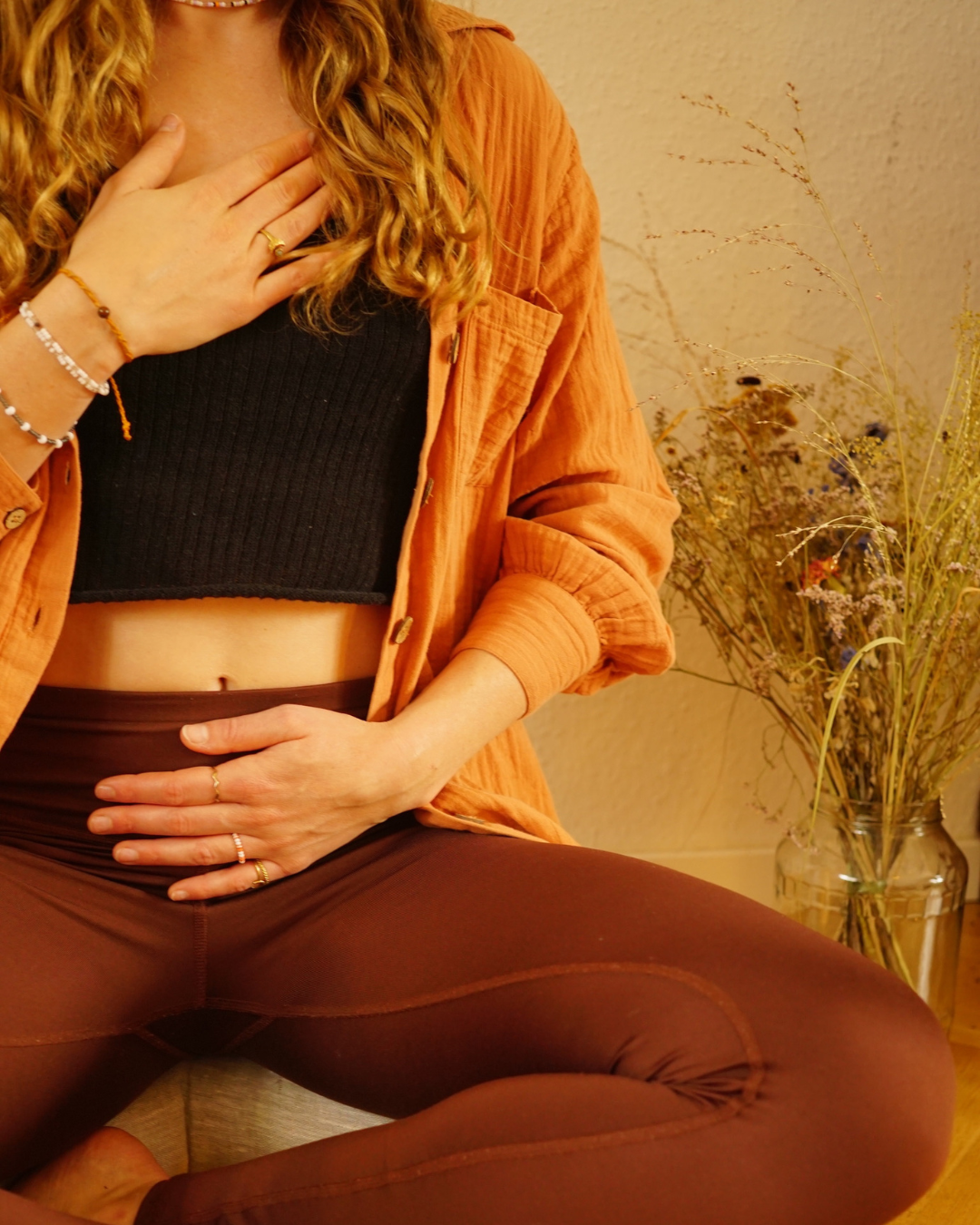 Eine junge Frau sitzt im Schneidersitz vor einer Vase mit getrockneten Blumen, trägt braune Leggings, ein schwarzes Oberteil, eine orange Jacke und hat lange, lockige Haare.