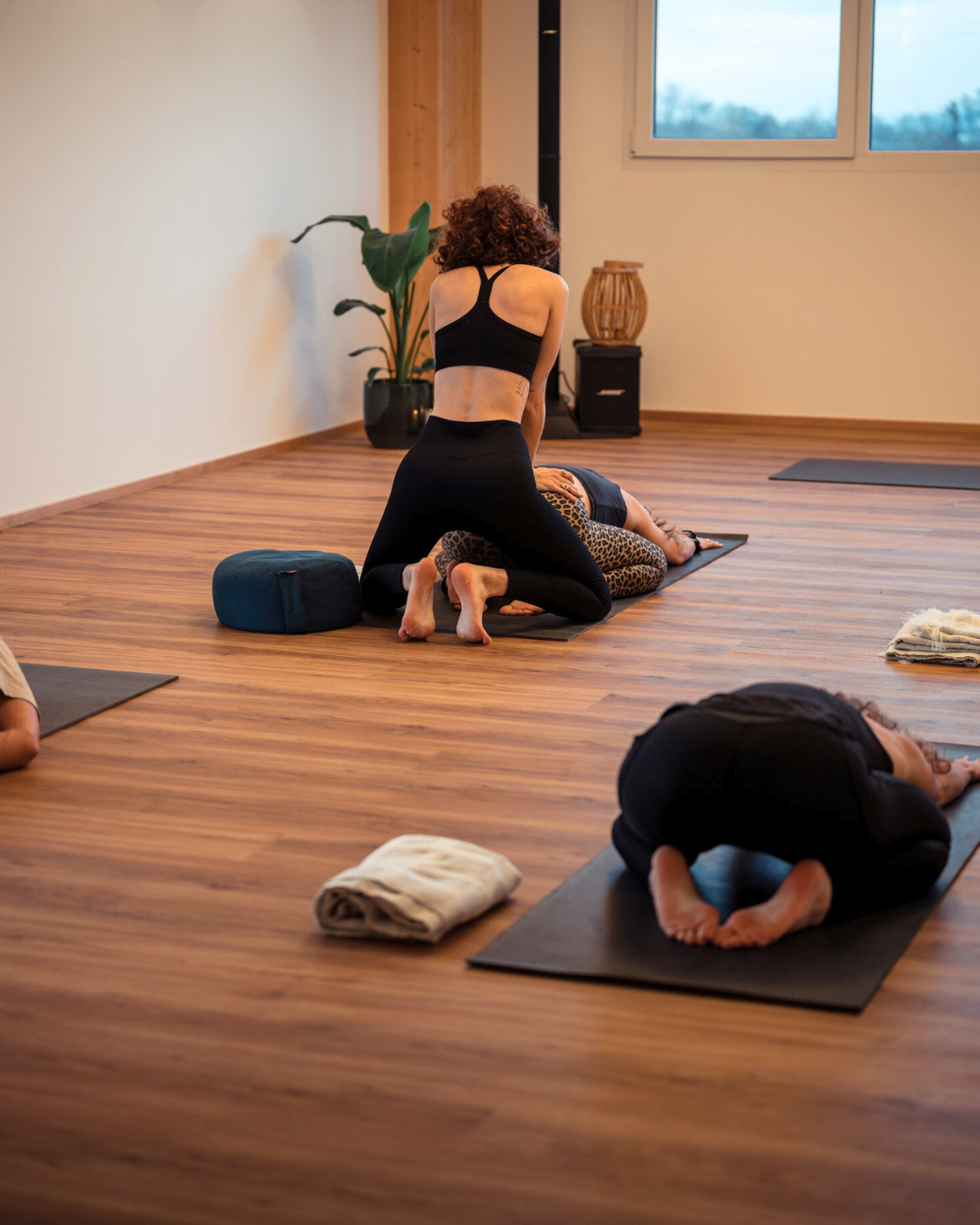 Eine Yoga-Lehrerin führt eine Yoga-Übung an, während die Schüler auf Yogamatten in einem hell erleuchteten Raum liegen.