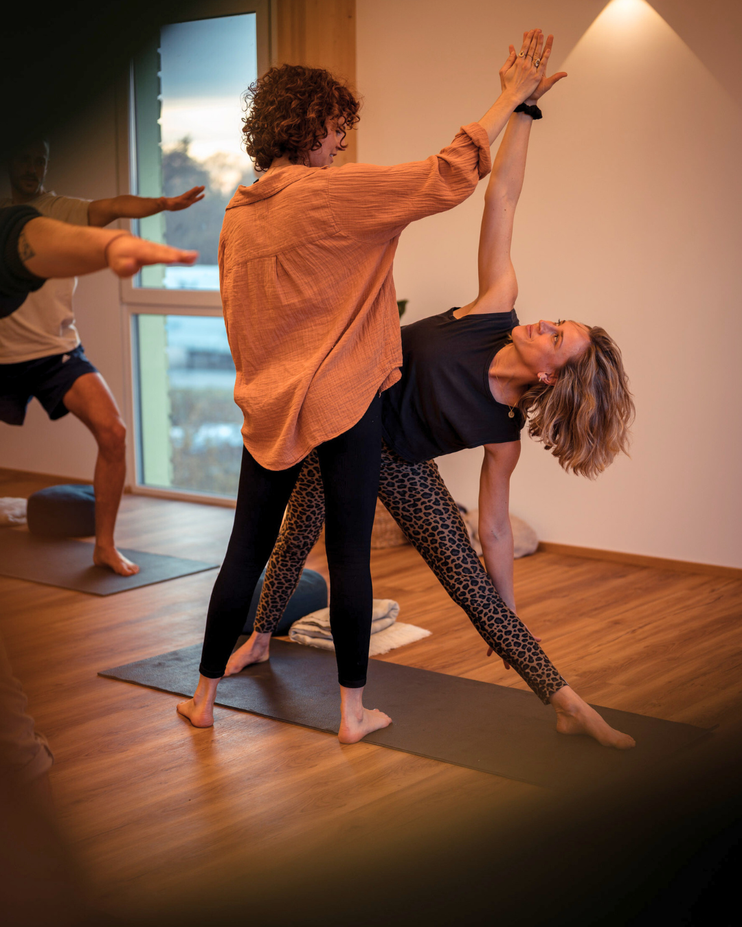 Zwei Frauen beim Yoga, eine hält den Arm der anderen nach oben, während eine dritte Person im Hintergrund eine ähnliche Yogapose einnimmt, in einem hellen Raum mit Holzfußboden.