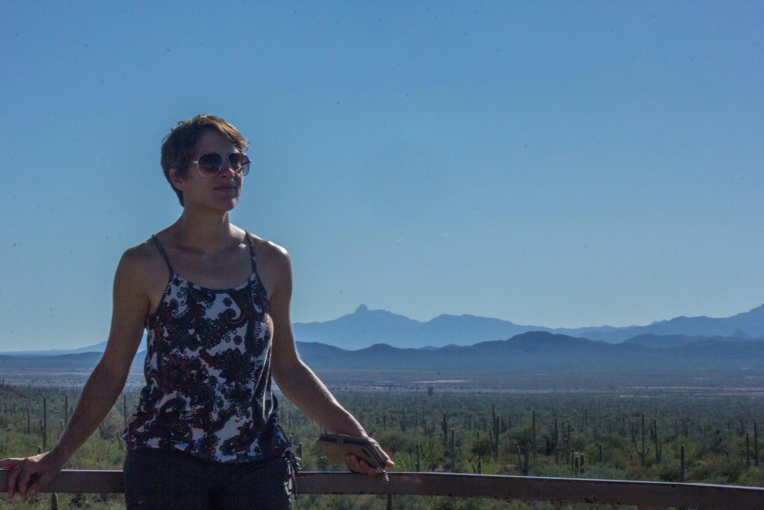 A woman wearing sunglasses and a patterned tank top walking outdoors with mountains and a blue sky in the background.