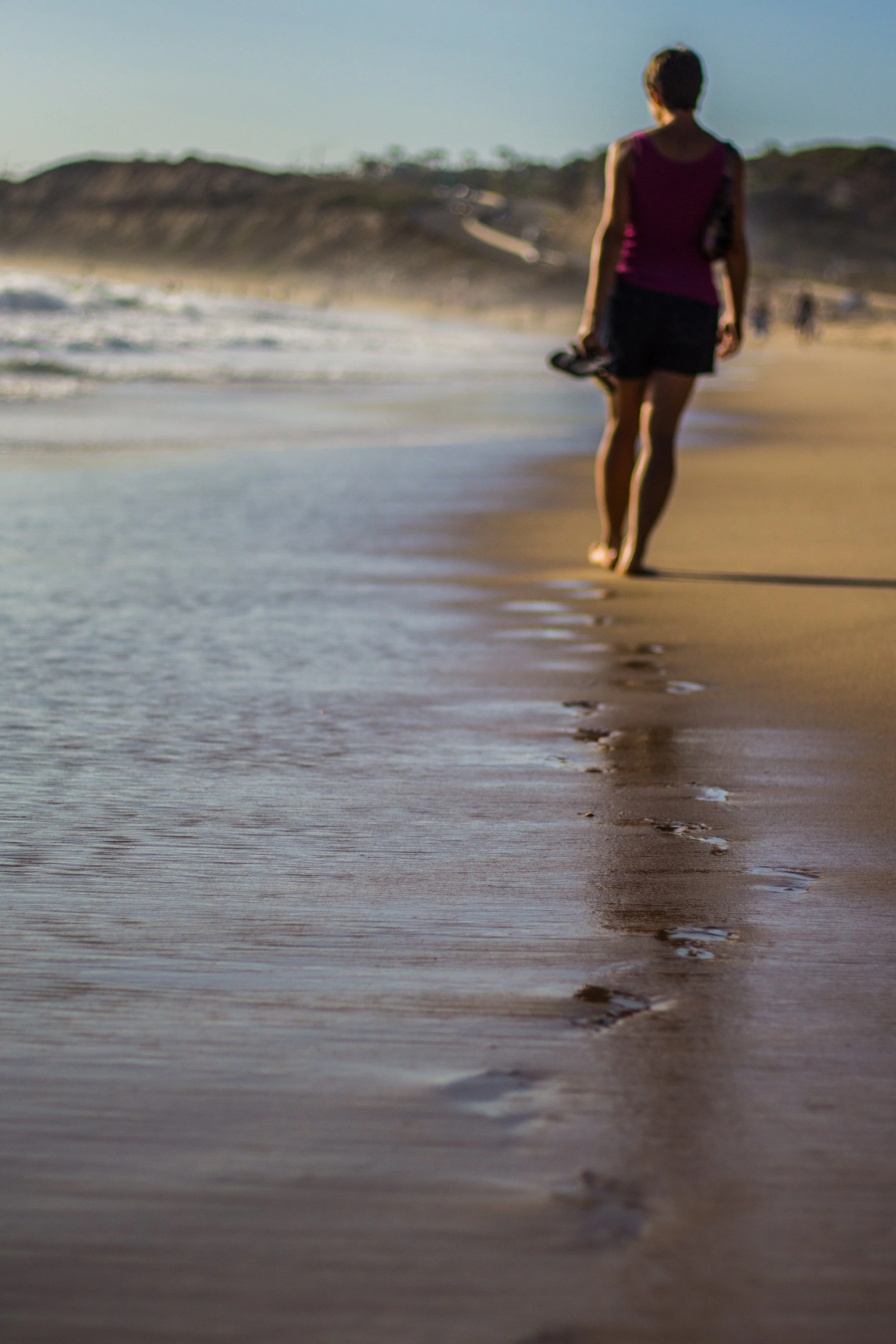 A woman walk along the shoreline holding flip-flops, with ocean waves and a distant cliff in the background, during sunset.