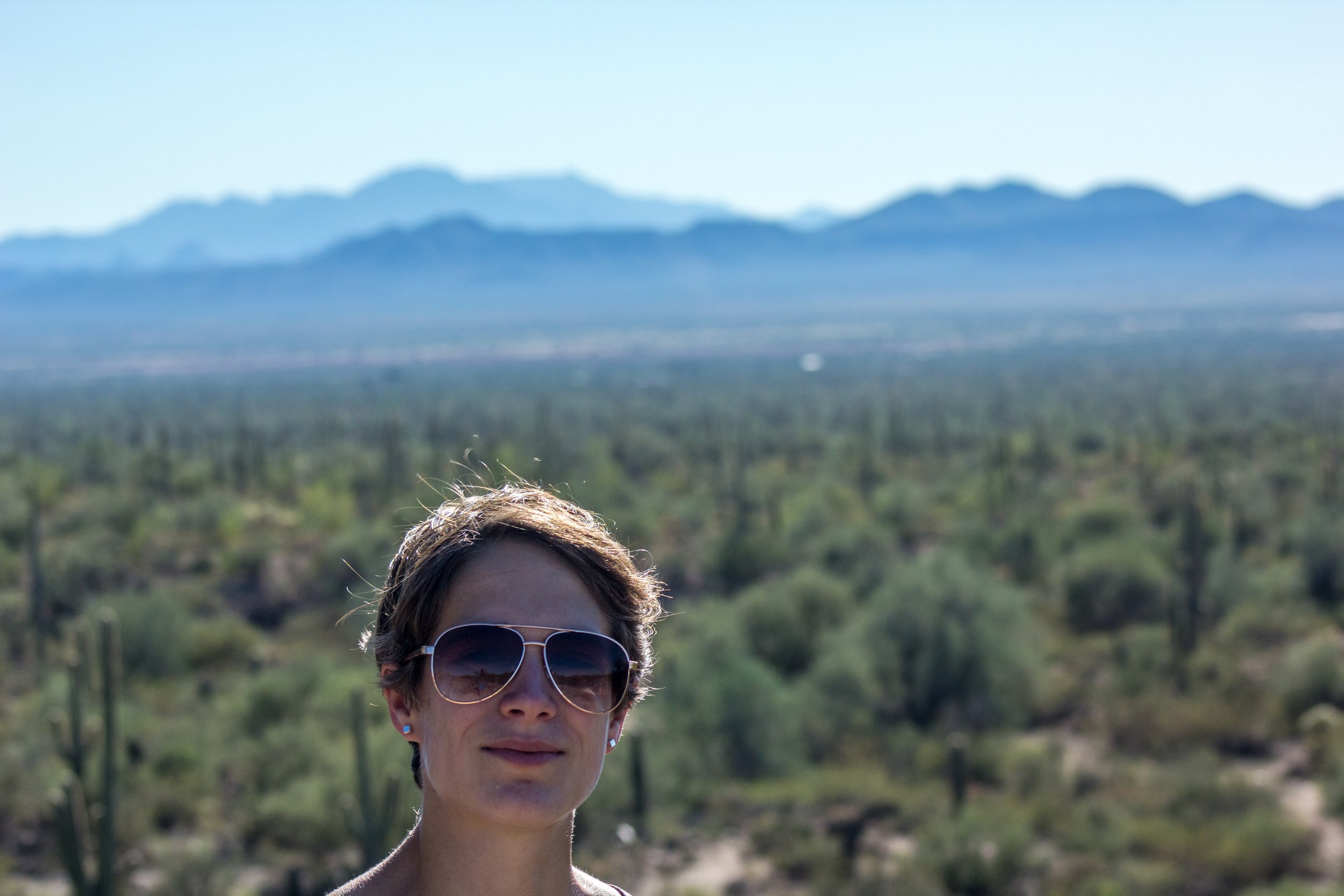 Close-up of a woman wearing sunglasses outdoors with mountains and desert vegetation in the background.