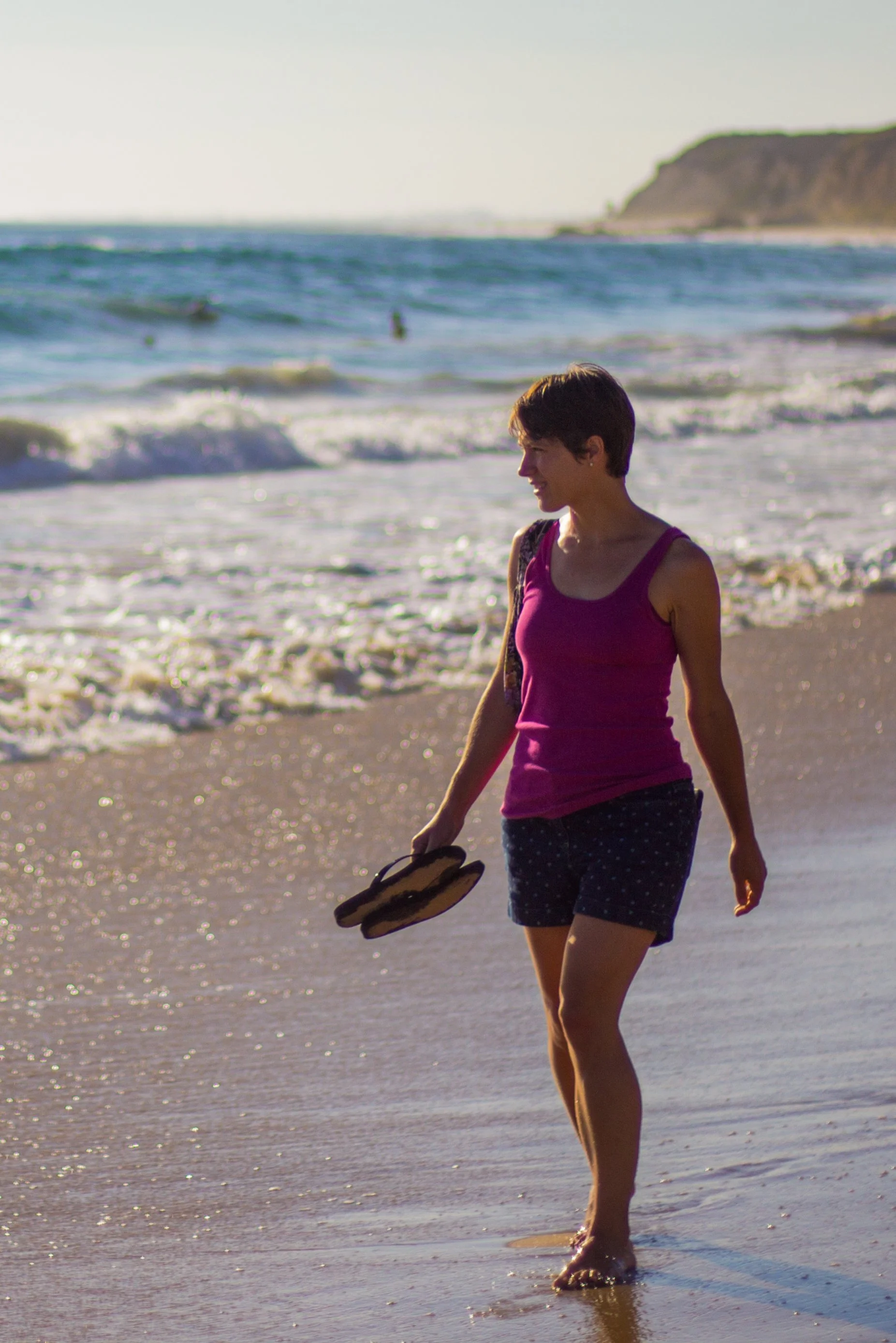 A woman in a purple tank top and navy polka dot shorts walking along the beach with her sandals in hand, waves in the background, and a hill in the distance.