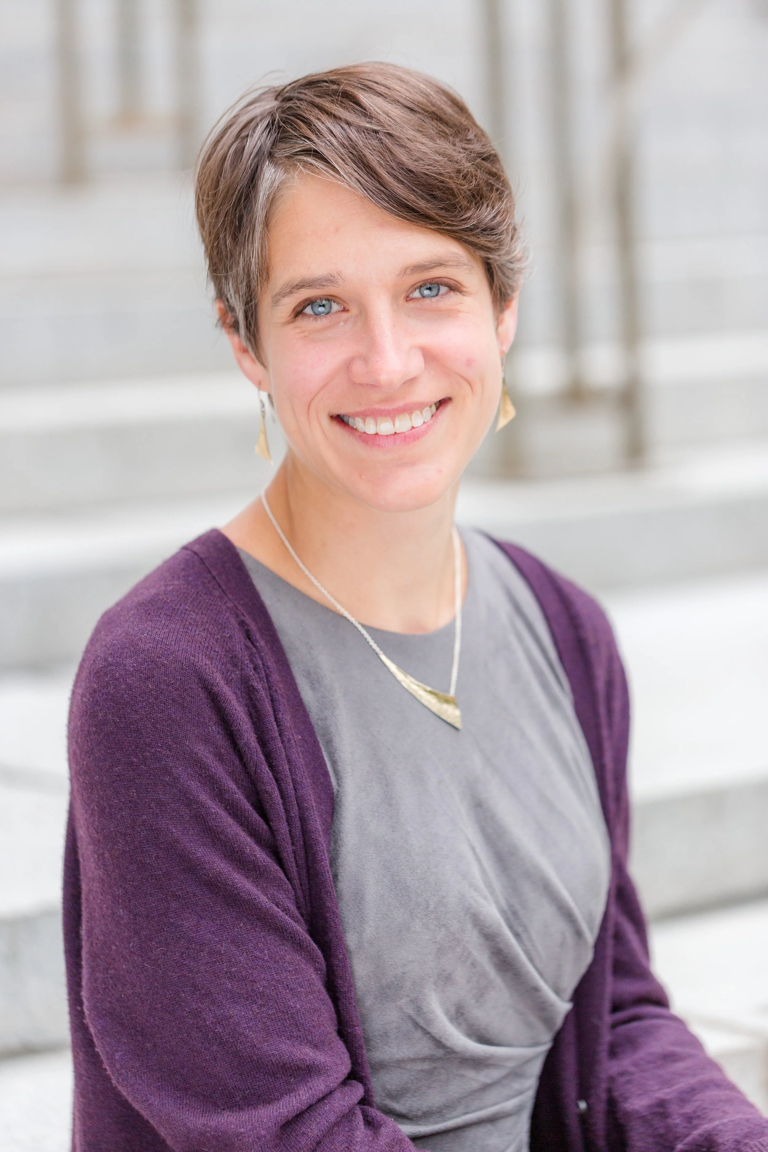 A woman with short brown hair, blue eyes, and light skin is smiling at the camera. She is wearing a gray shirt, a purple cardigan, and gold earrings, with a silver necklace. She is sitting outdoors on a staircase with a blurred background.