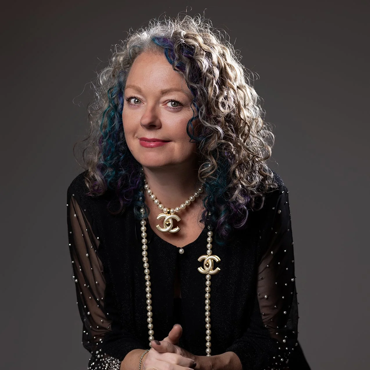 A woman with curly, multi-colored hair (blonde with purple, blue, and green highlights) wearing a black top with pearl and Chanel jewelry, including layered necklaces and a large Chanel logo pendant, posed against a gray background.