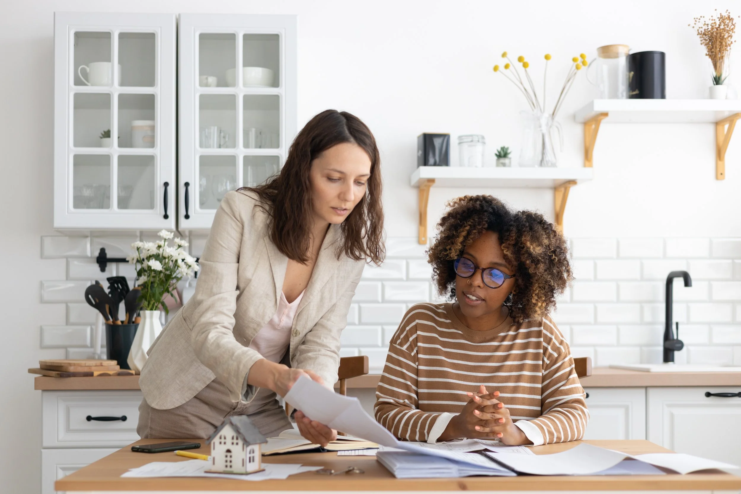 Two women, one with brown hair and the other with curly hair and glasses, are working together at a kitchen table, reviewing papers and documents. The kitchen has white cabinetry, shelves, and decorations including a small house model and a vase with white flowers.
