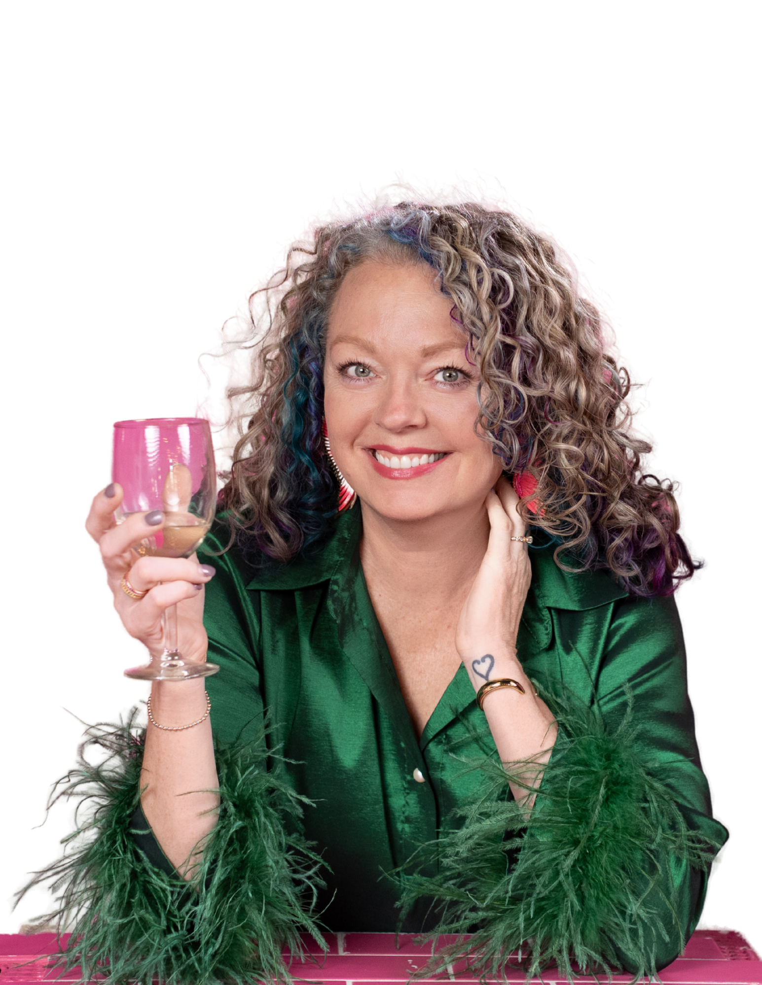 Woman with curly blonde hair with blue and purple streaks, smiling, holding a pink glass, wearing a green velvet top, surrounded by green foliage, possibly marijuana leaves, in front of a pink table.