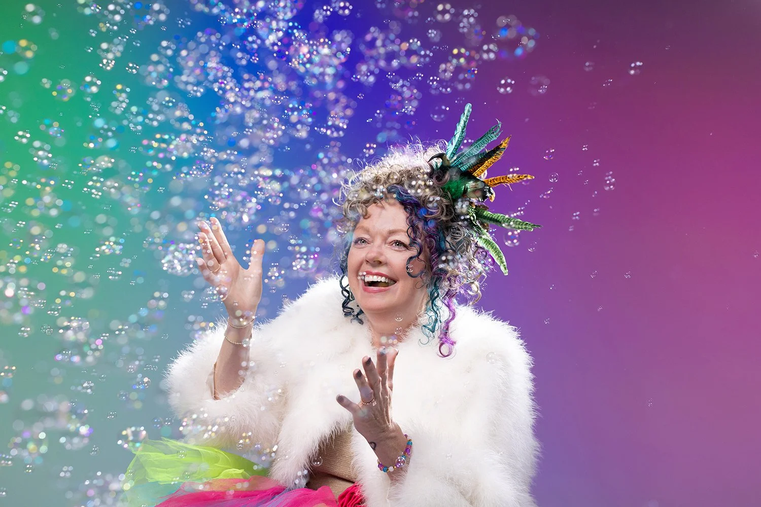 A joyful woman with curly, multicolored hair decorated with feathers, wearing a white furry coat and a pink and green tutu, is tossing glitter in the air against a rainbow-colored background.
