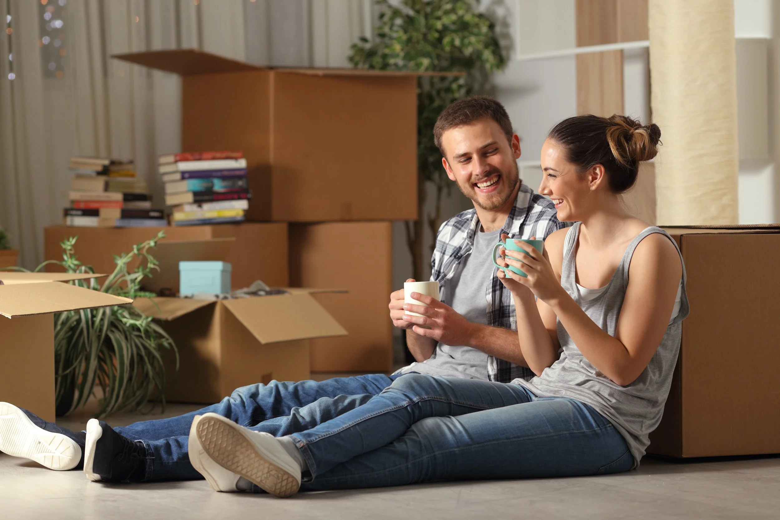 Two people sitting on the floor amidst moving boxes and packing supplies, smiling and holding coffee mugs, in a living room with a stack of books and a potted plant in the background.