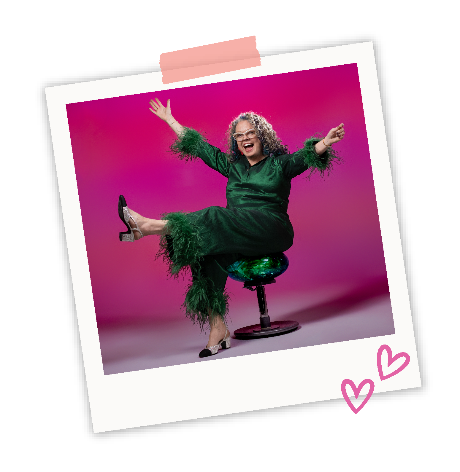 Lin Reynolds sitting on a stool with a pink background, wearing a dark green satin outfit with feathered sleeves and cuffs, high heels, and smiling with arms raised.