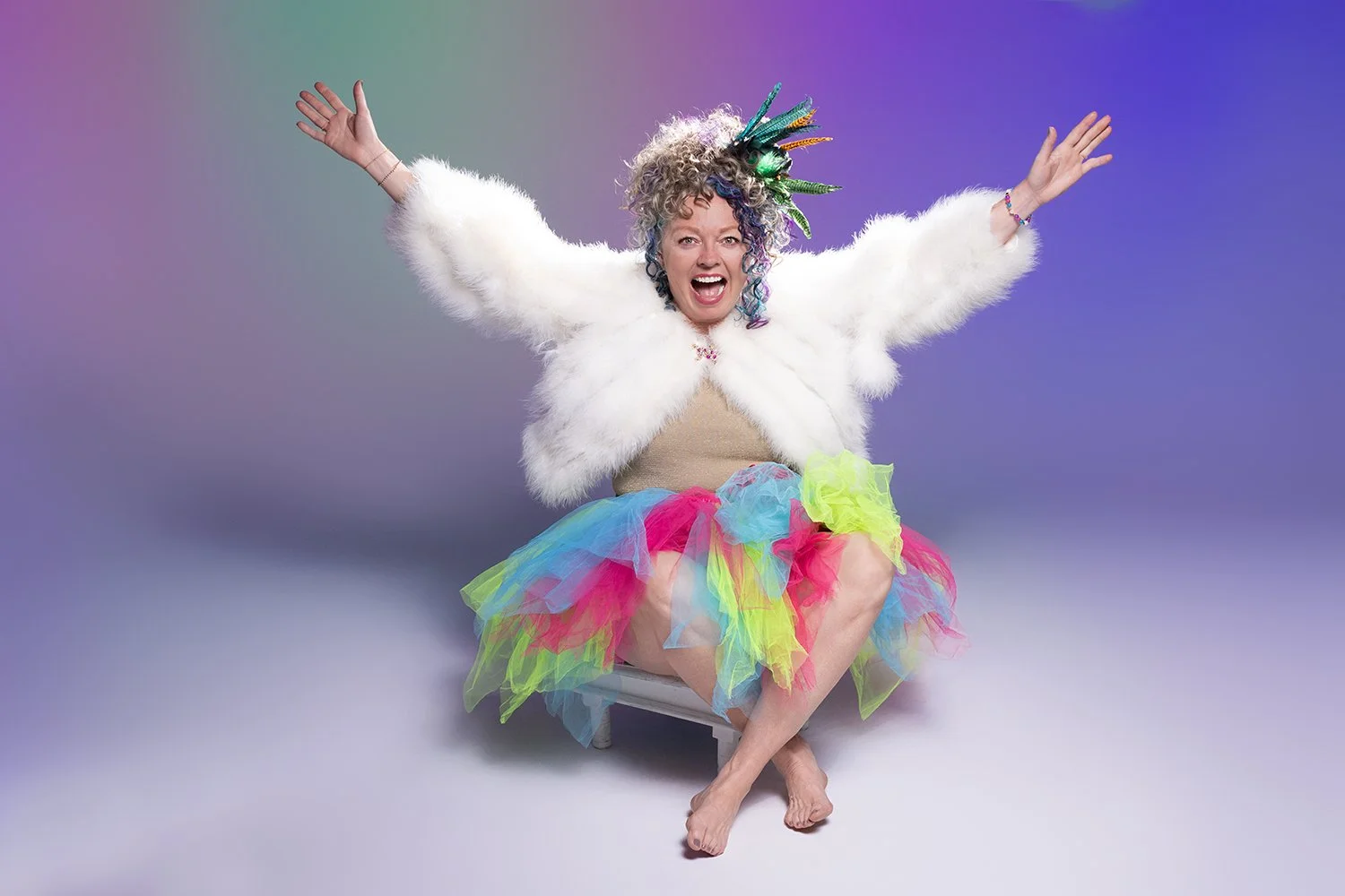 Woman with curly hair wearing a colorful tutu, white fur jacket, and a feathered headdress, sitting with arms raised and smiling in front of a rainbow gradient background.