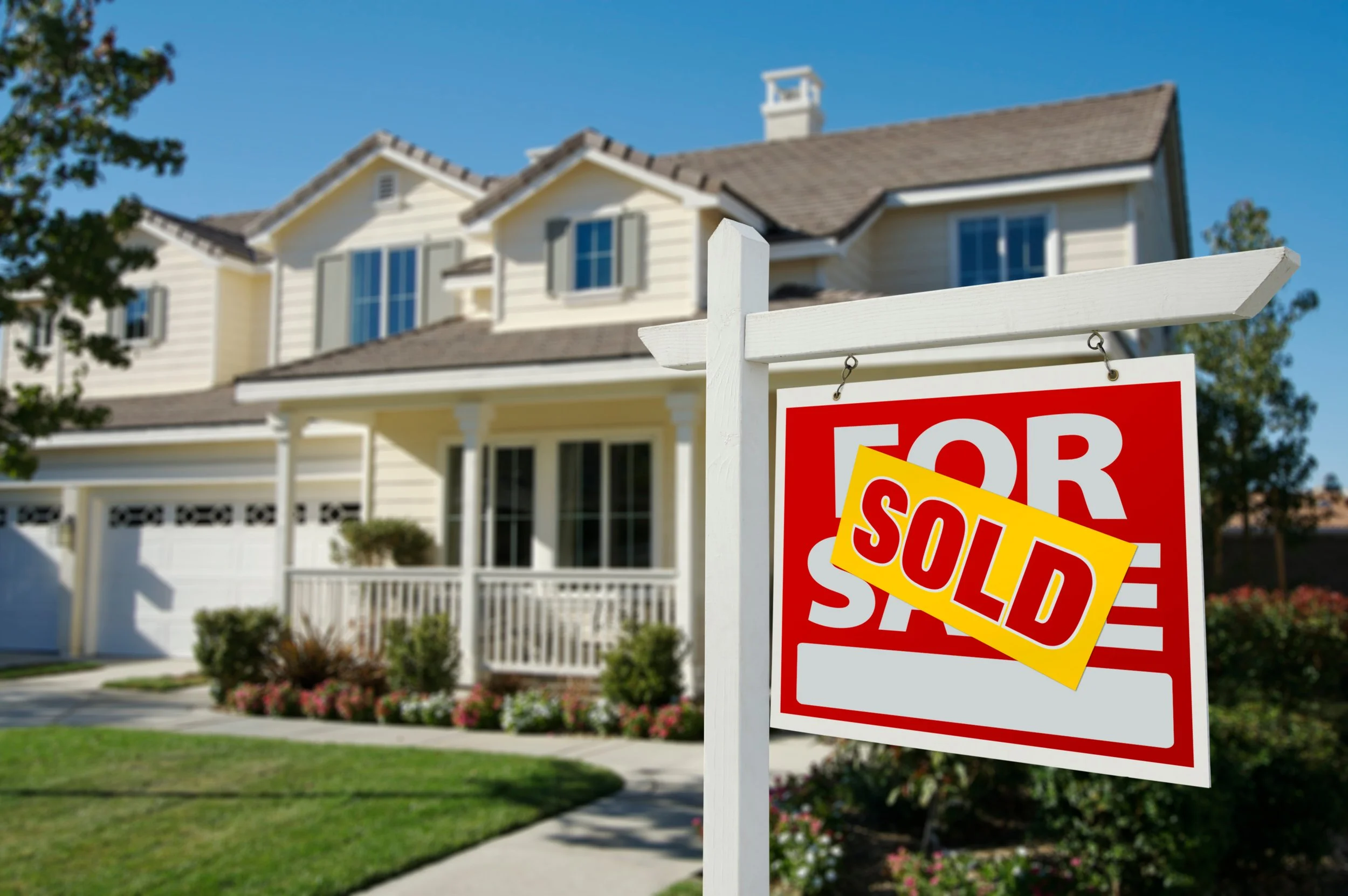 A house with a "For Sale" sign in front of it, partially covered by a "Sold" sign. The house is cream-colored with gray shutters and a brown shingled roof.