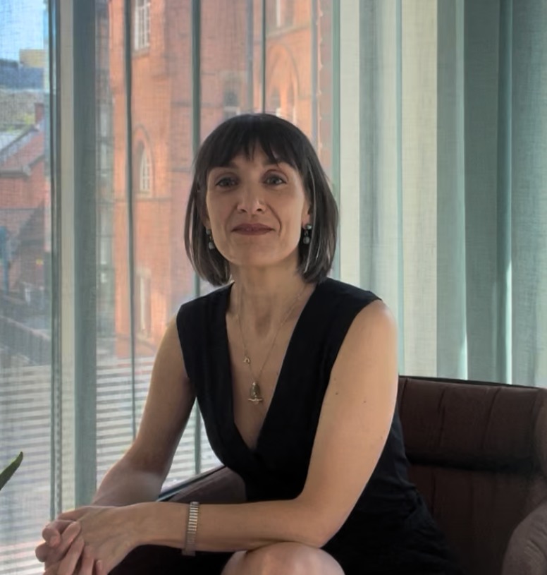 A woman with short dark hair, wearing a black sleeveless dress, sitting indoors near large windows with city buildings in the background.