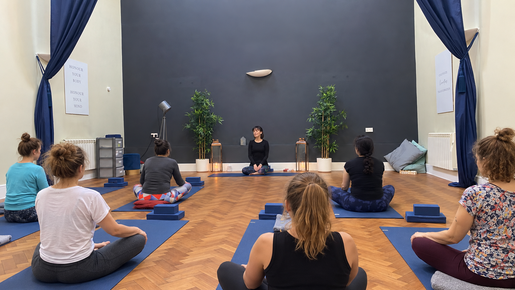A group of women participating in a yoga or meditation class in a spacious, well-lit room with dark blue curtains on the sides, wooden flooring, and a black accent wall. The instructor sits cross-legged in front, with plants and lanterns behind her.