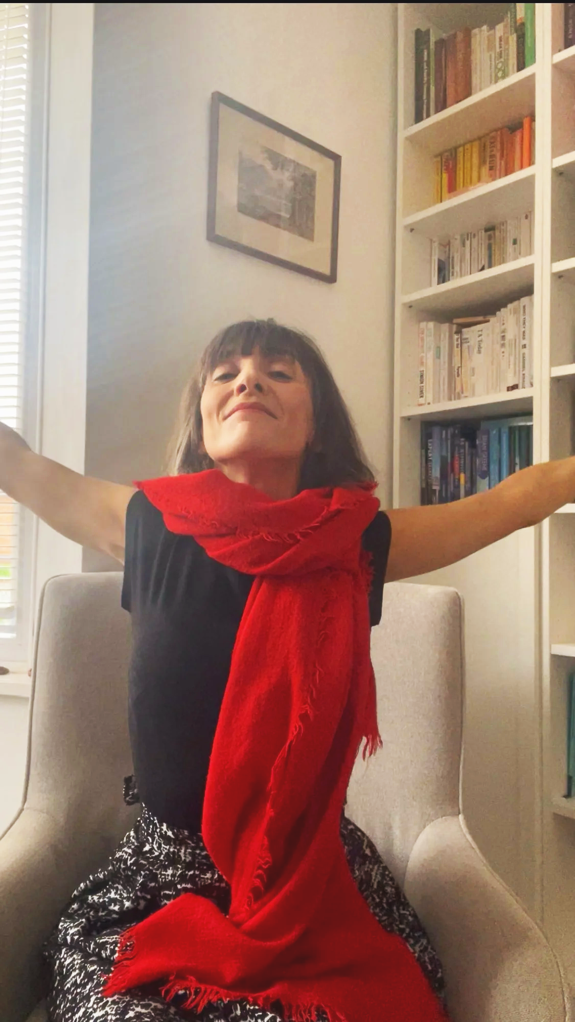 Person with arms outstretched sitting on a chair in front of a white bookshelf, wearing a red scarf and black top.