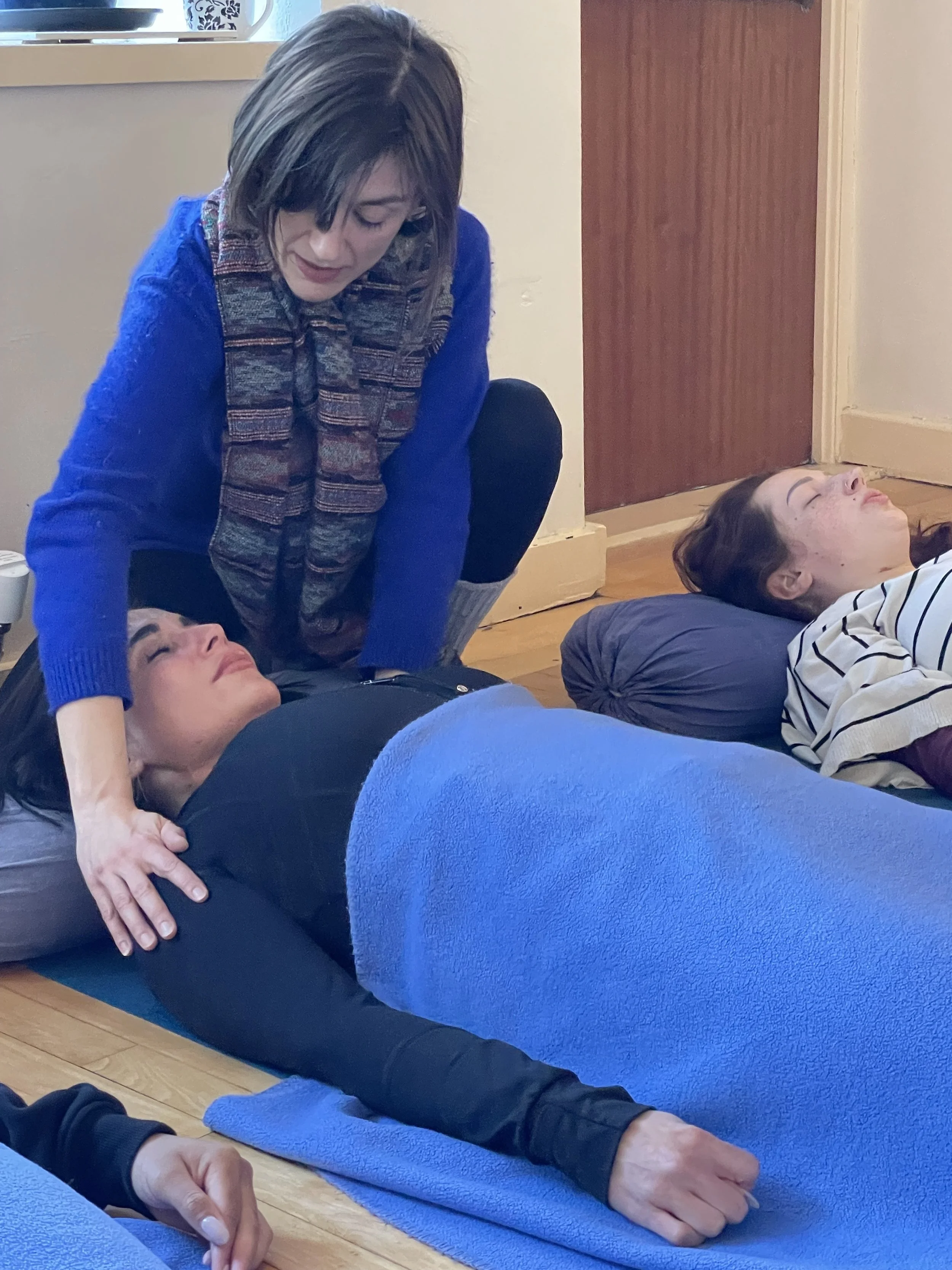 A woman is lying face down on a mat with her eyes closed, with another woman kneeling beside her, placing her hand on her back, possibly gently pressing or massaging.