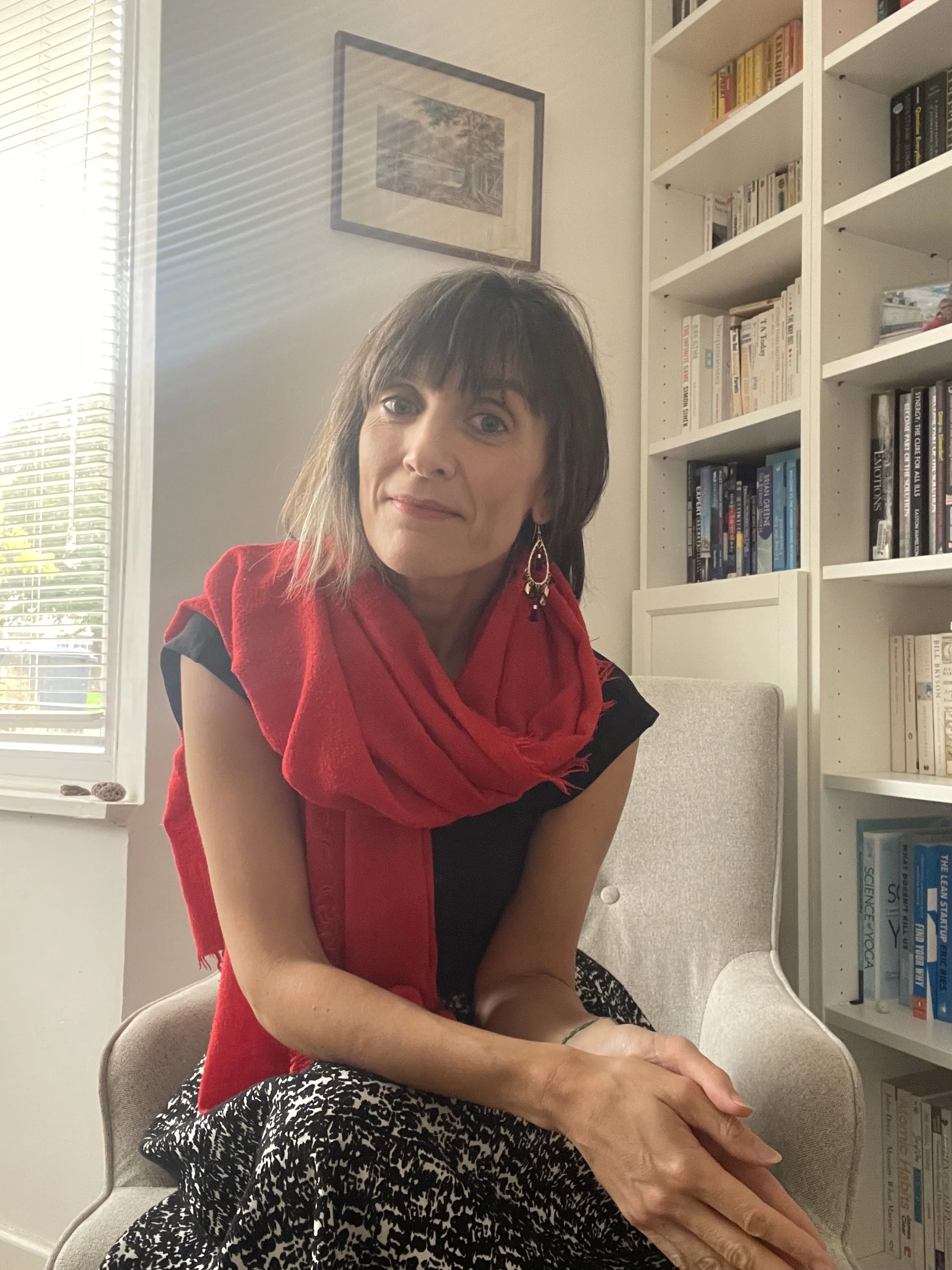 A woman with brown hair and bangs, wearing a red scarf and black top, sitting on a beige armchair in a room with white bookshelves filled with books in the background, and beside a window with white blinds.