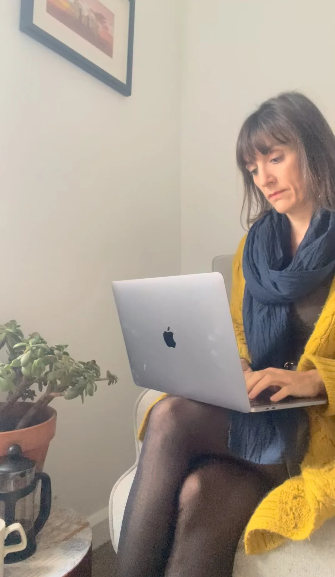 A woman with dark hair and bangs, wearing a yellow sweater and black tights, sitting on a chair while working on a silver MacBook laptop. There is a potted plant on a small table next to her, and a wall with a framed picture hanging on it.