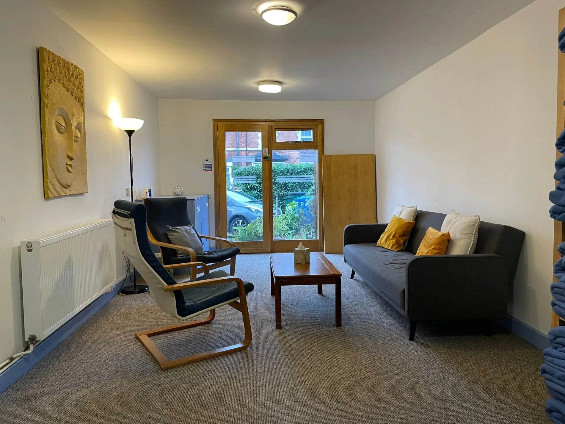 A waiting room with a gray couch with white and yellow pillows, two black armchairs, a wooden coffee table with a tissue box, a tall standing lamp, a wall decoration of a face sculpture, a radiator, and a door with glass panels showing a parking lot outside.