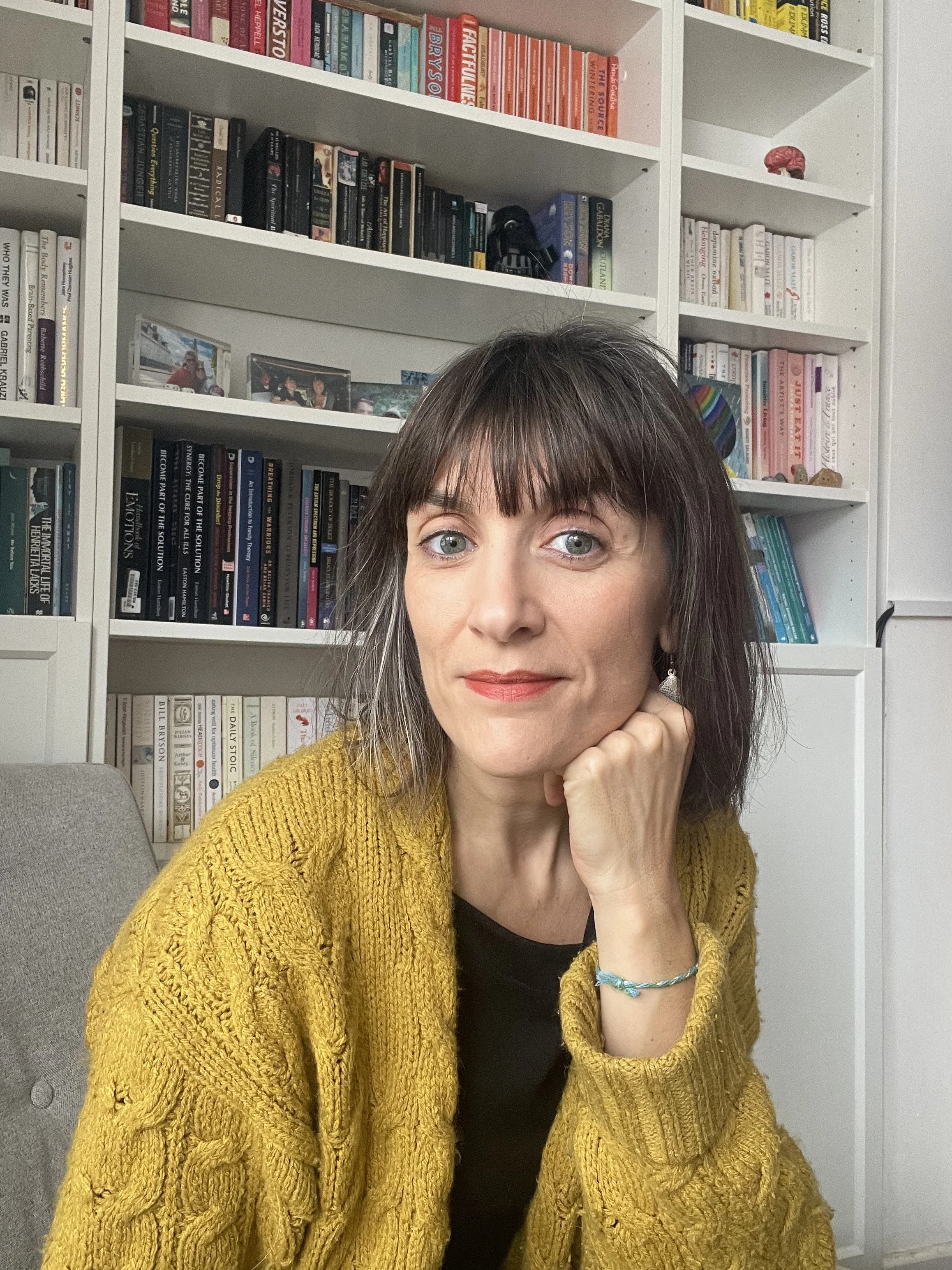 A woman with dark hair and light eyes, wearing a yellow cable knit sweater, sitting in front of bookshelves filled with colorful books, resting her chin on her hand, smiling gently.