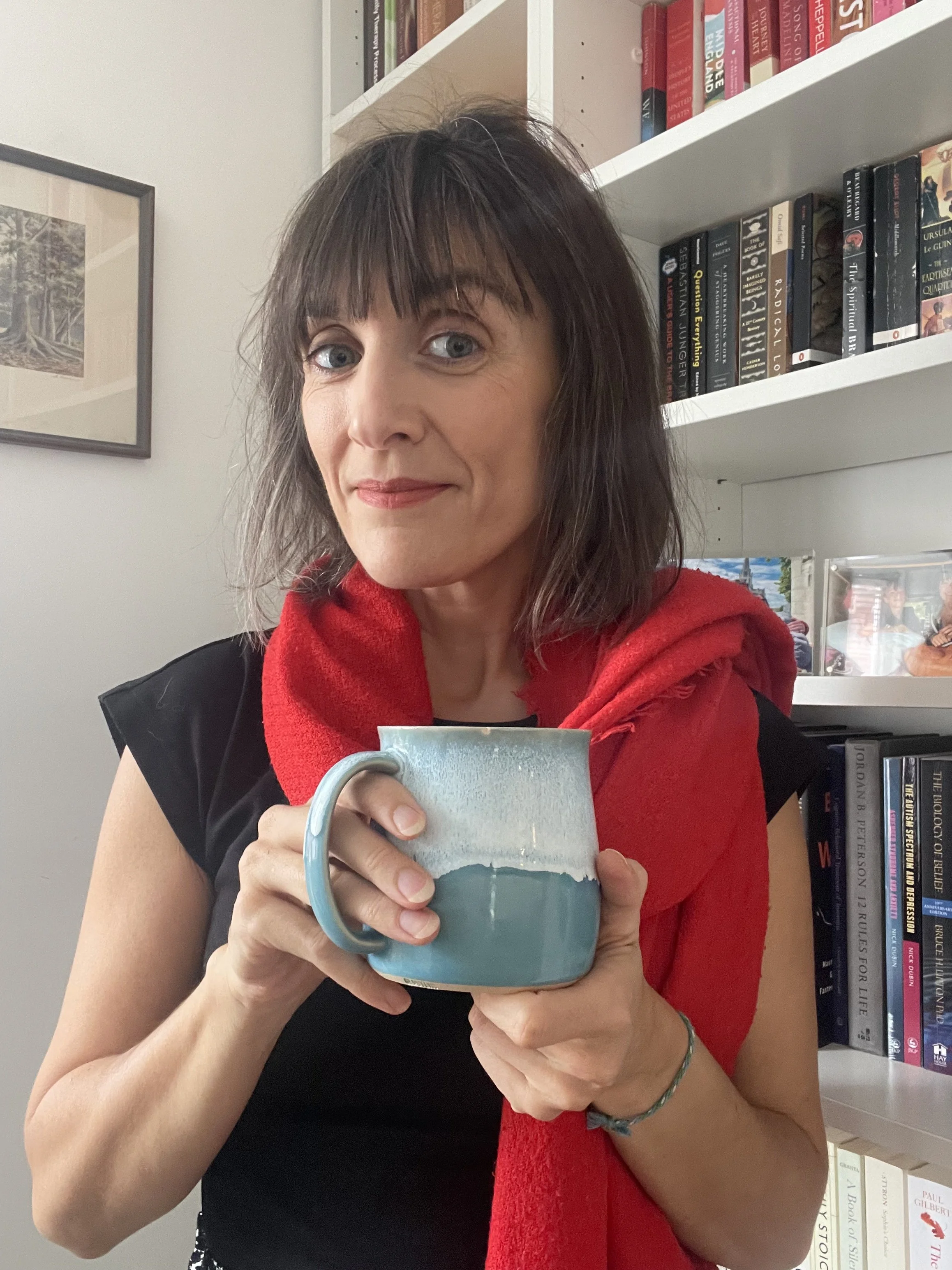 A woman with dark hair and blue eyes holding a blue and beige ceramic mug, wearing a black top and a red scarf, standing in front of a bookshelf filled with books and a framed photo on the wall.