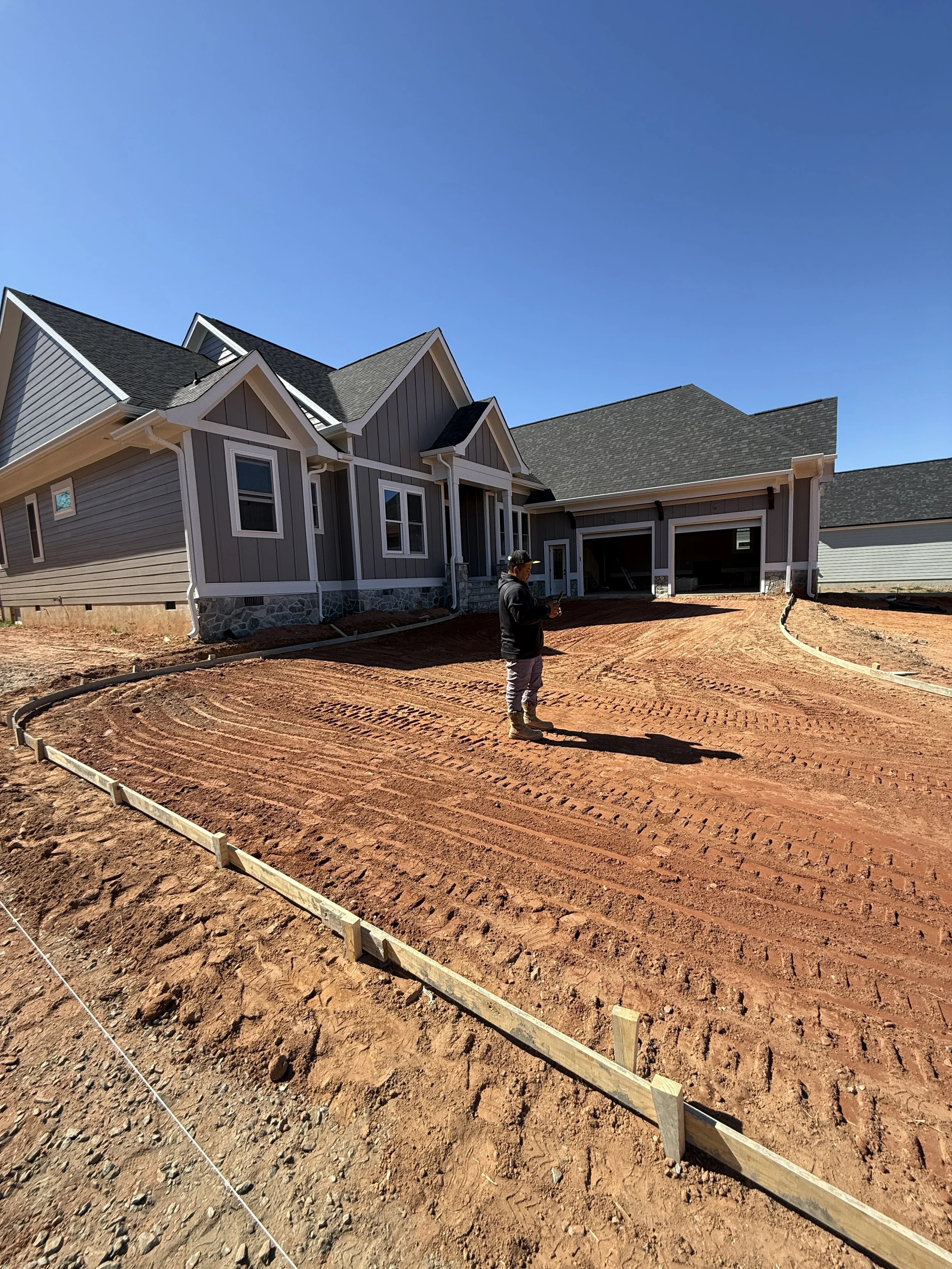 A newly constructed house with gray siding and a dark roof, a person standing on dirt ground, and a clear blue sky in the background.