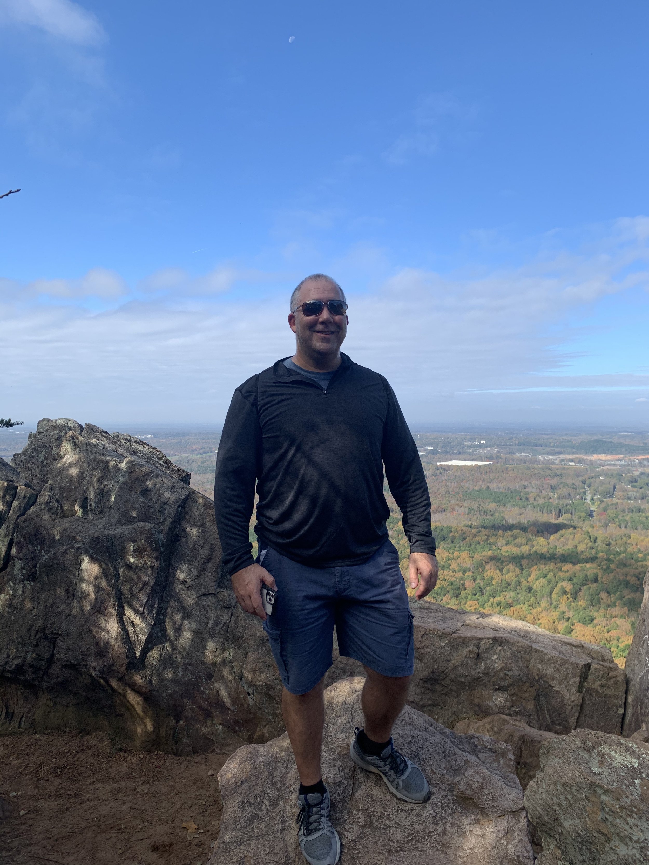 A man wearing sunglasses, a black jacket, gray shorts, and sneakers standing on a large rock at a high elevation. He is smiling and holding a smartphone, with a scenic landscape of trees and fields in the background under a partly cloudy sky with the moon visible.