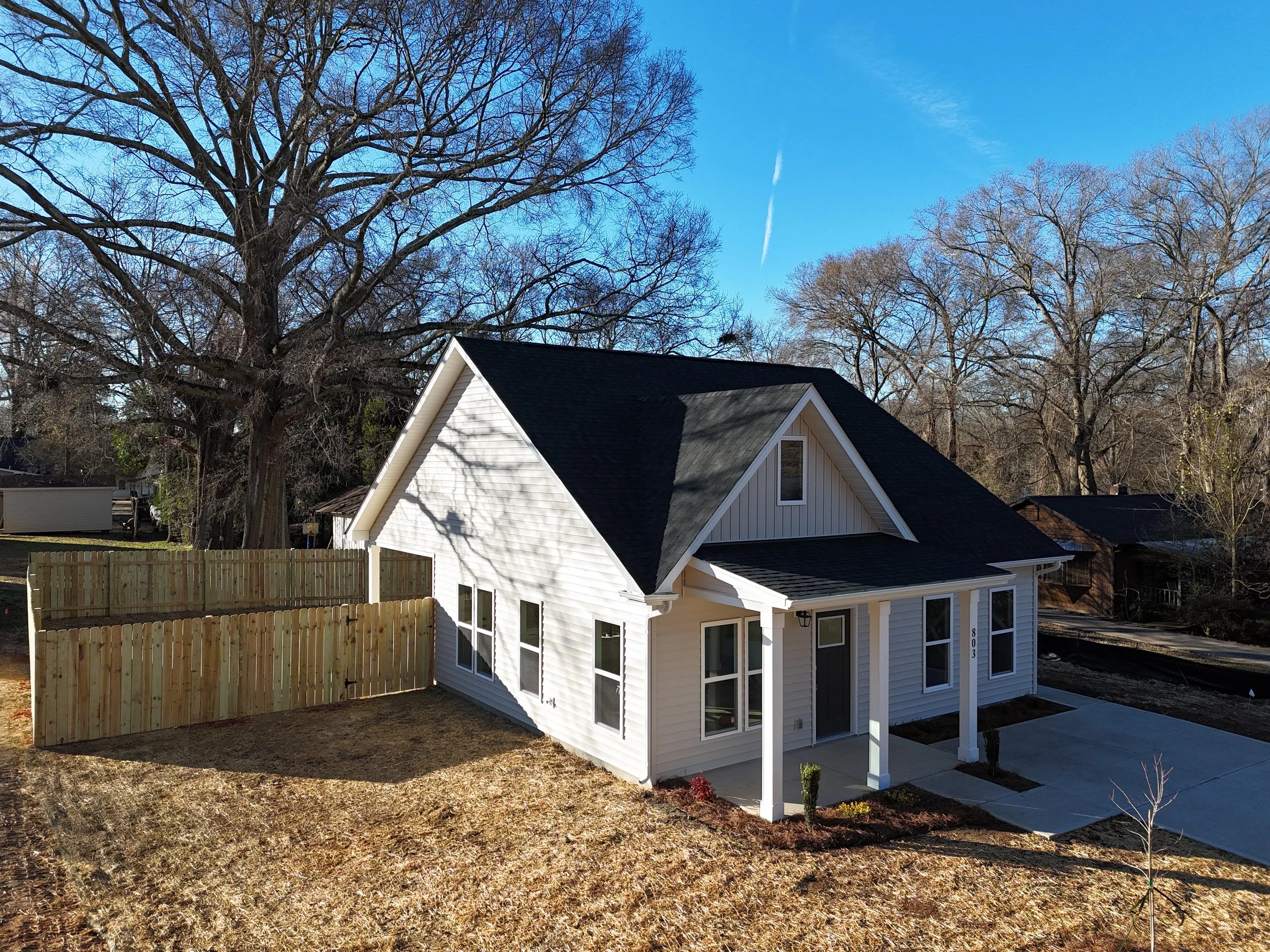 A white house with a dark roof and multiple windows features a small front porch and a sloped roof. The yard is partly grass and dirt, surrounded by a wooden fence. Tall, leafless trees and a blue sky with thin clouds are in the background.