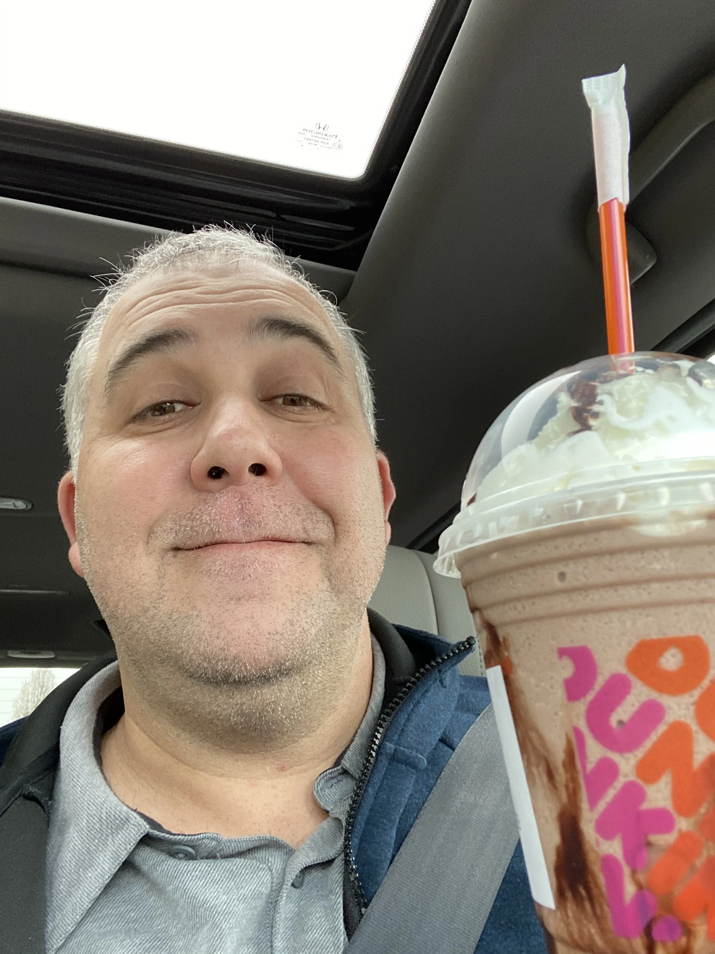 Half of a middle-aged man's face with gray hair and light skin, sitting in a car, holding a large milkshake with whipped cream and caramel drizzle, with a strab in the car.
