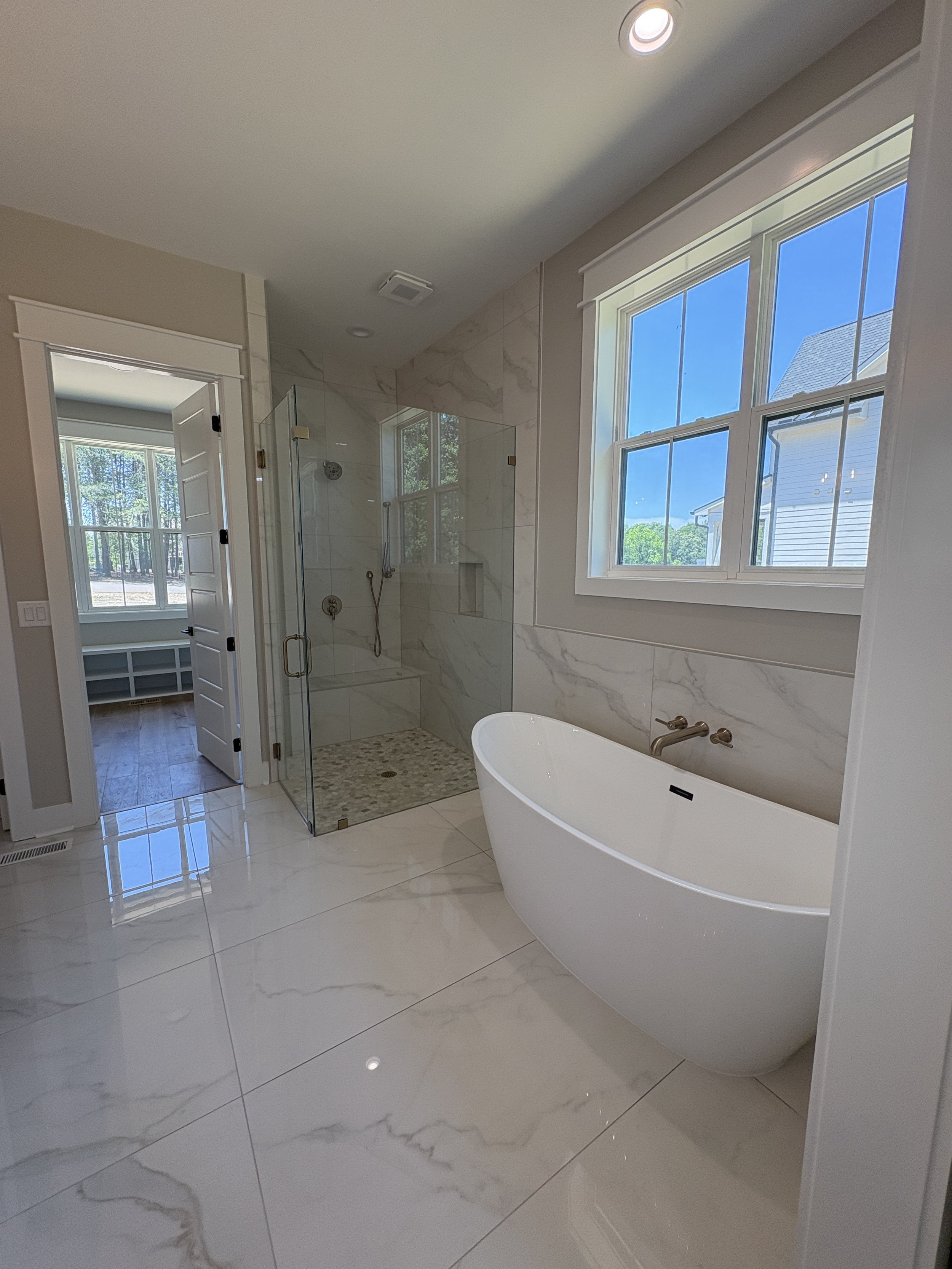 Modern bathroom with walk-in shower, frameless glass enclosure, gold hardware, freestanding bathtub, large window with a view of trees and neighboring house, and tile flooring and walls.