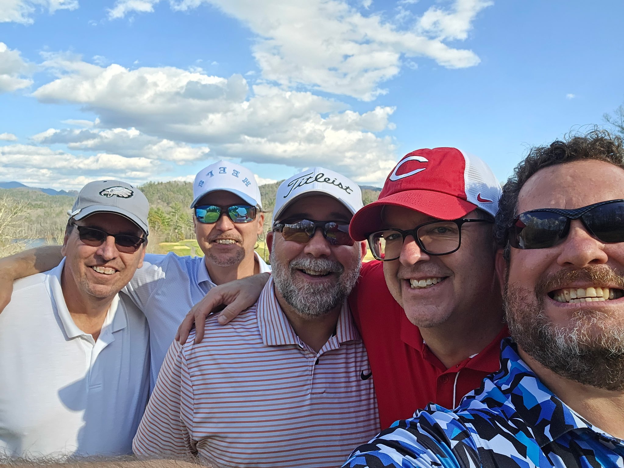 A group of five men in casual golf attire, smiling and taking a selfie outdoors at a golf course, with mountains and a blue sky with white clouds in the background.