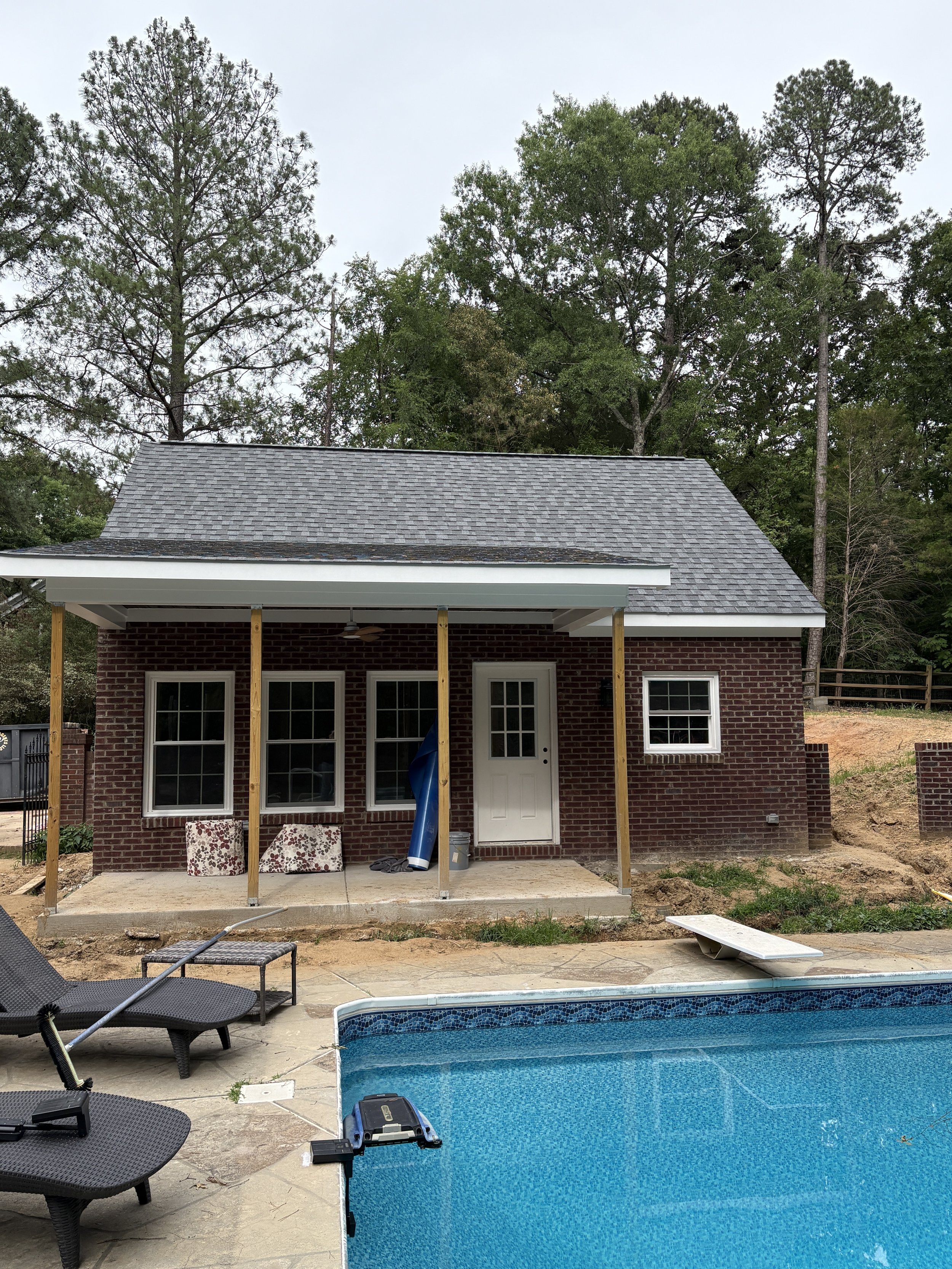 A backyard scene with a swimming pool, patio chairs, and a brick house with a new porch under construction. The house has a gray shingle roof, white trim, and several windows. The background features tall trees and a cloudy sky.
